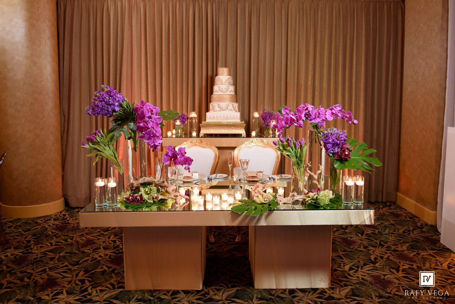 Elegant wedding dessert table with tiered white cake, purple orchids, candles, and floral arrangements in a grand ballroom.