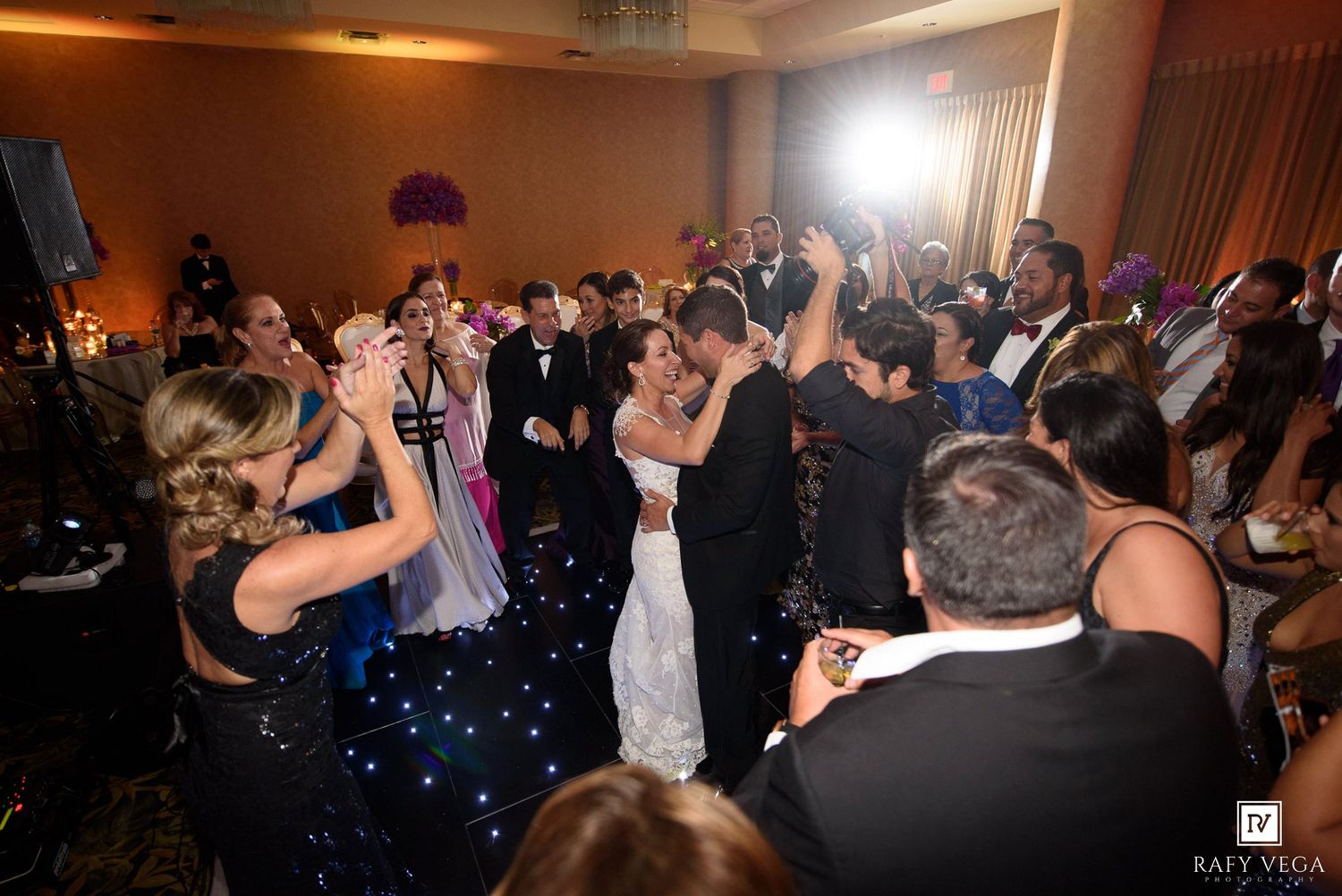Bride and groom share first dance surrounded by guests at elegant wedding reception with dark star-lit dance floor.