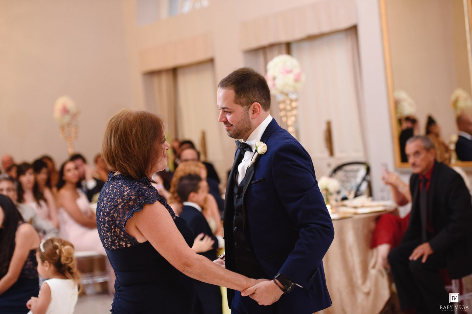 Groom in navy suit shares a dance with his mother during elegant wedding reception.