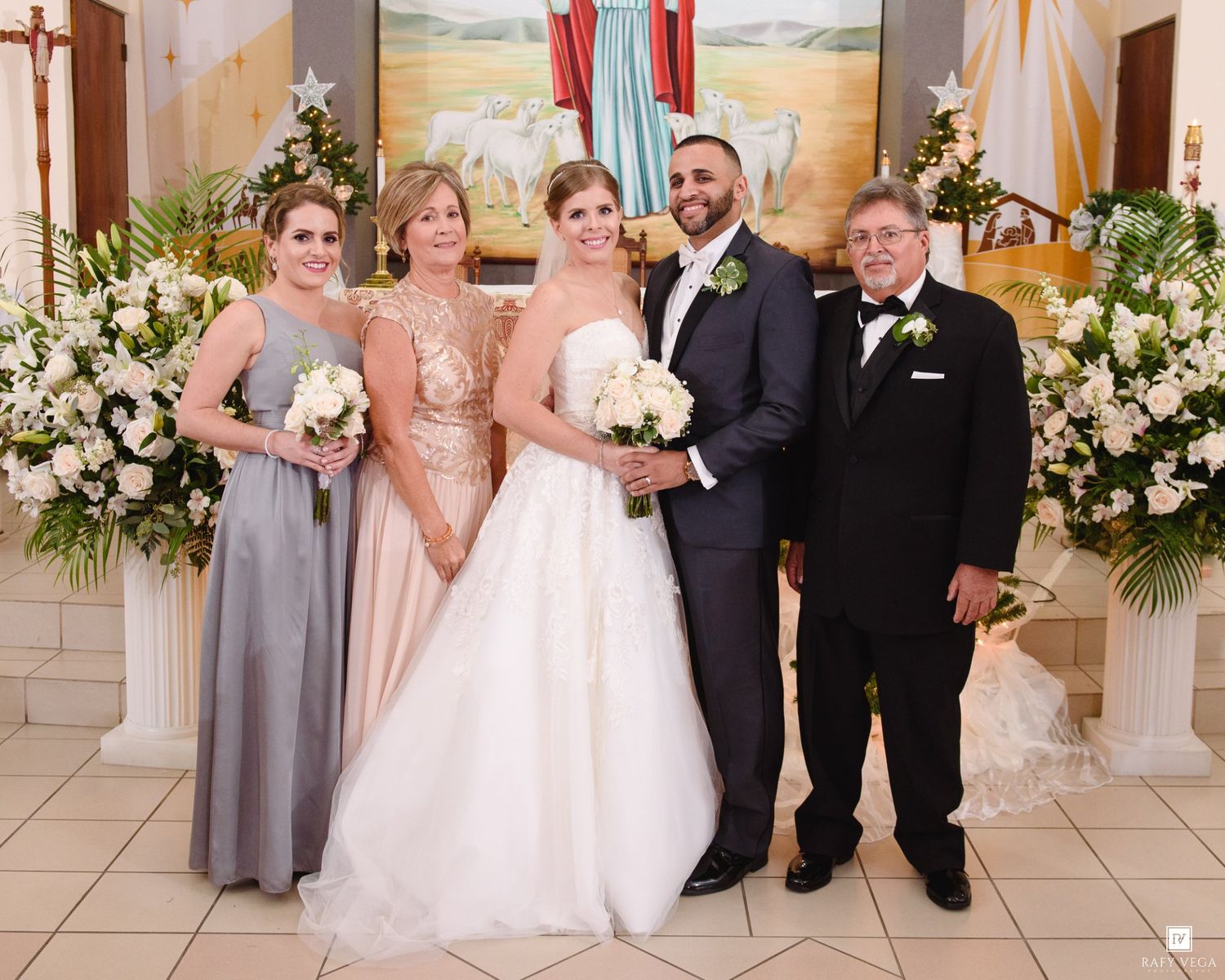 Wedding party poses with bride and groom at church altar surrounded by white floral arrangements.