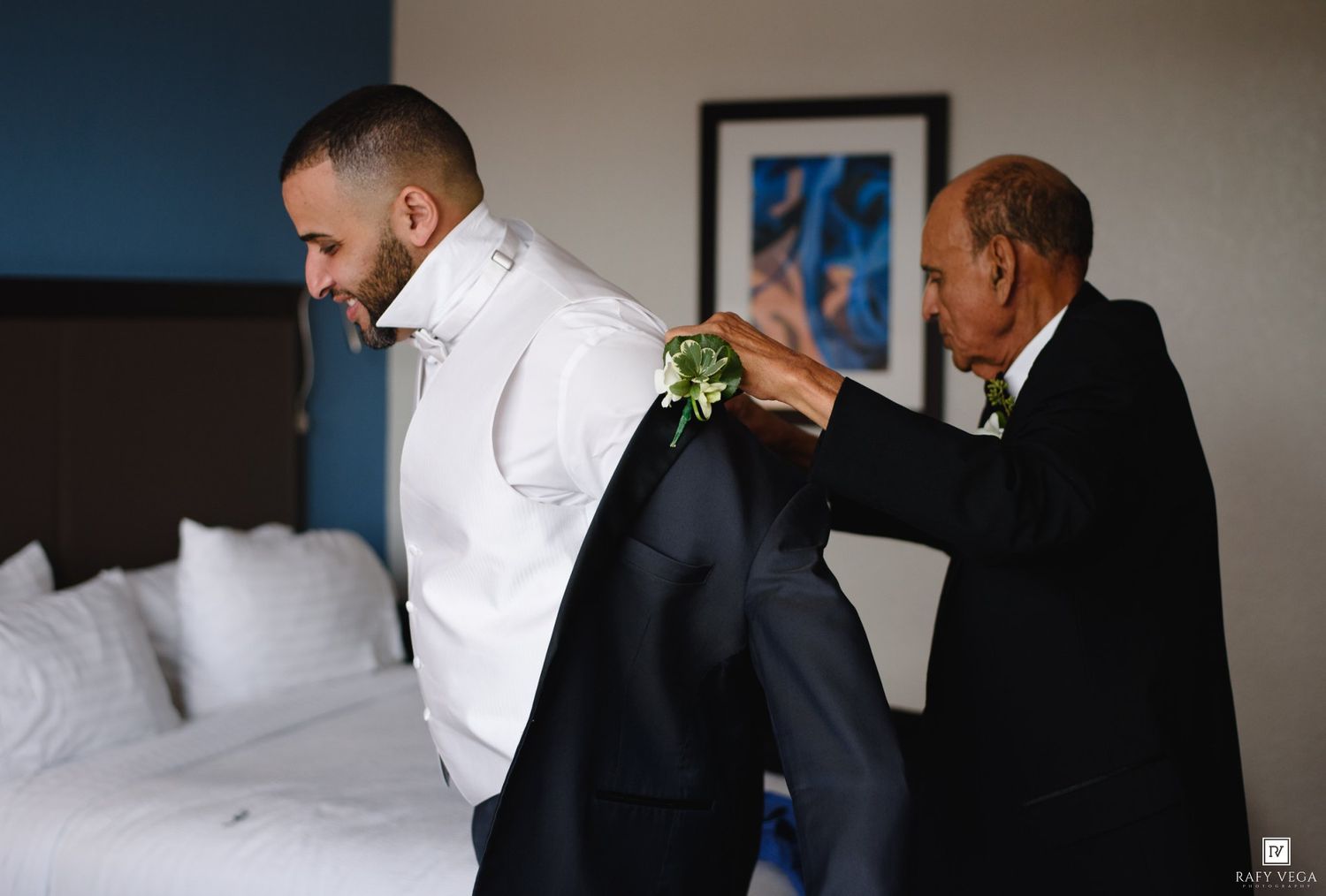 Groom in navy suit gets boutonniere pinned by older man in hotel room before wedding ceremony.