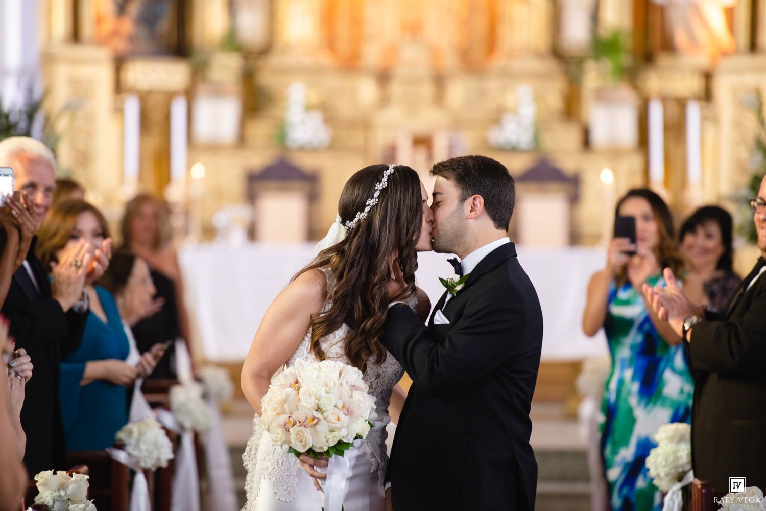 Bride and groom share first kiss at church altar during wedding ceremony with guests applauding in background.
