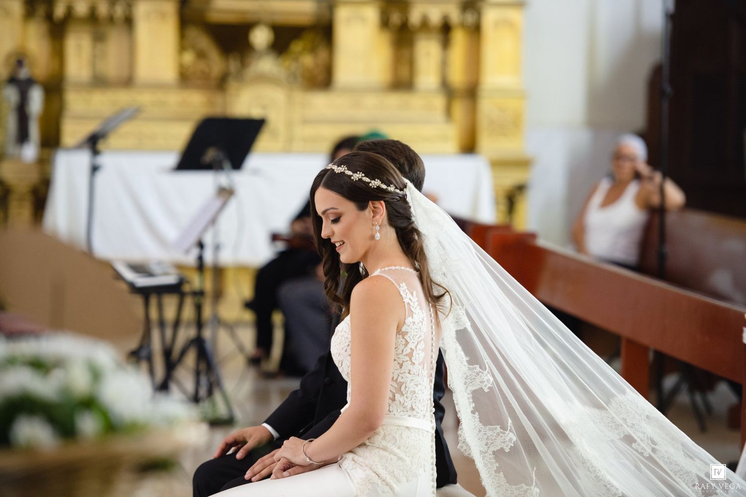 Beautiful bride in white lace gown and veil seated at church altar during wedding ceremony.