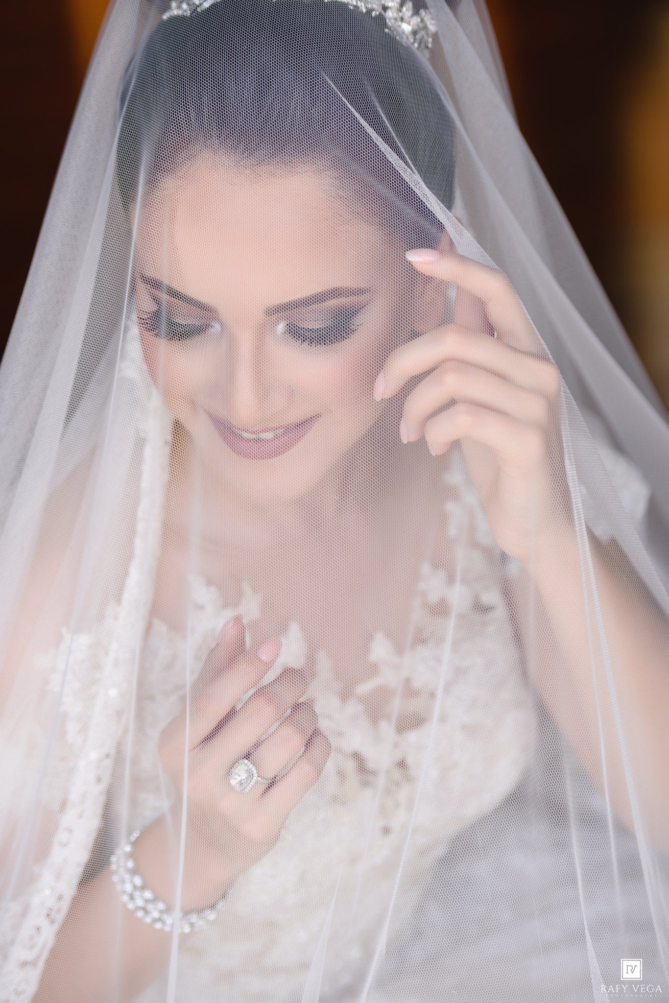 Elegant bride in lace gown and veil, wearing pearl bracelet and diamond ring, smiling softly with bridal crown.