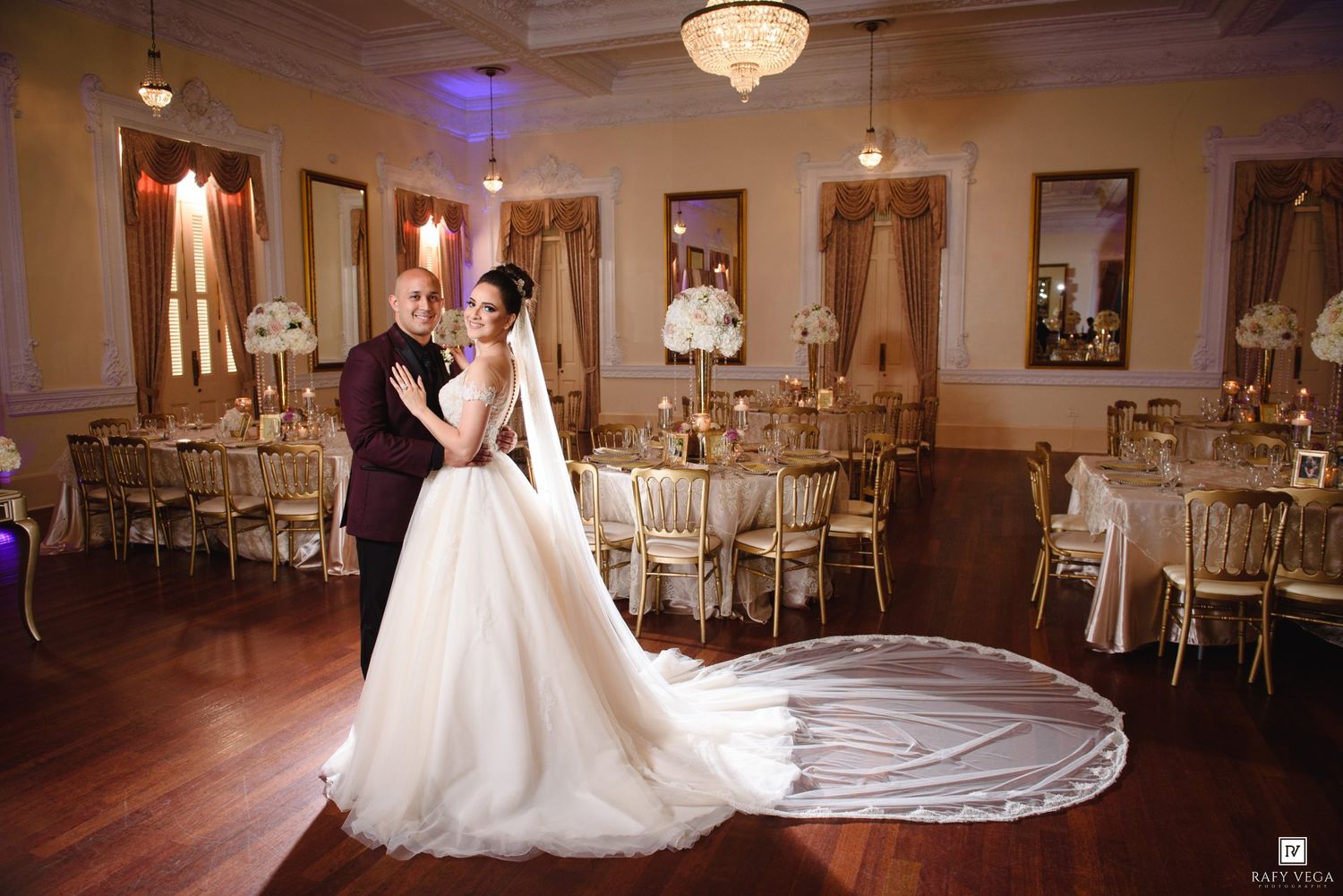 Bride and groom pose in an elegant ballroom with gold chairs and chandelier lighting at their wedding reception.