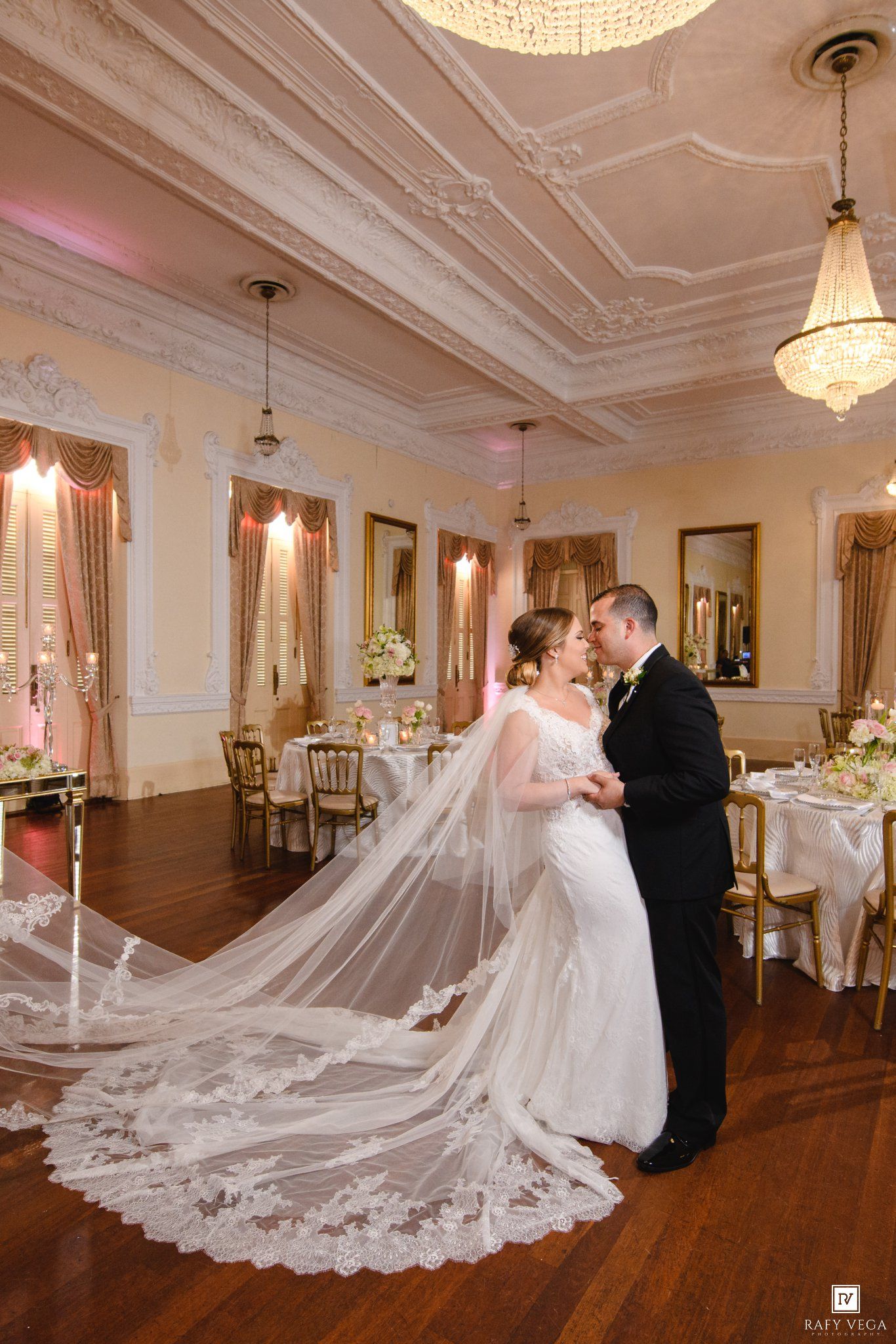 Bride and groom share a romantic kiss in an elegant ballroom with ornate ceilings, chandeliers, and a flowing lace cathedral veil.