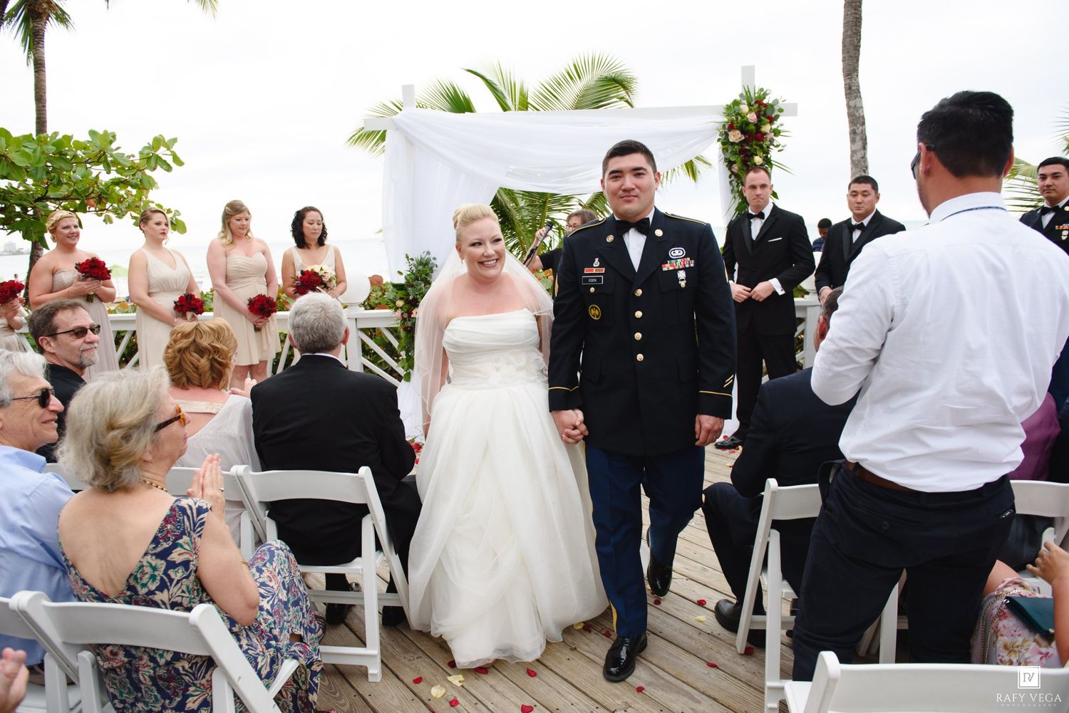 Military groom in dress uniform walks bride down aisle at outdoor beach wedding ceremony with guests seated on white chairs.