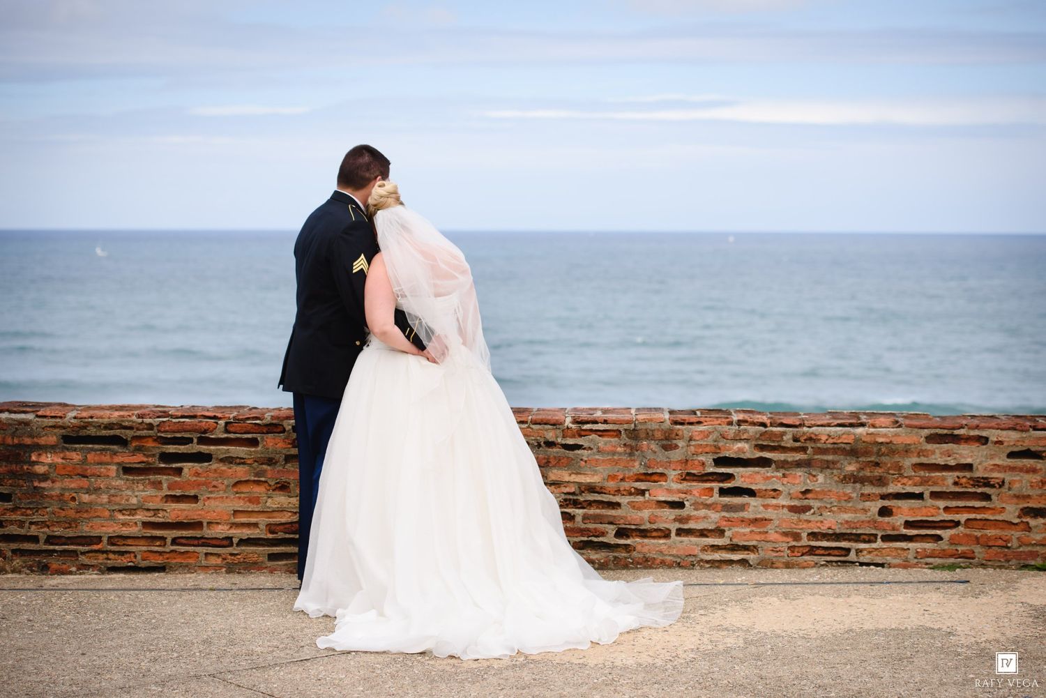 Bride and groom share romantic moment overlooking the ocean at a brick seaside wall on their wedding day.