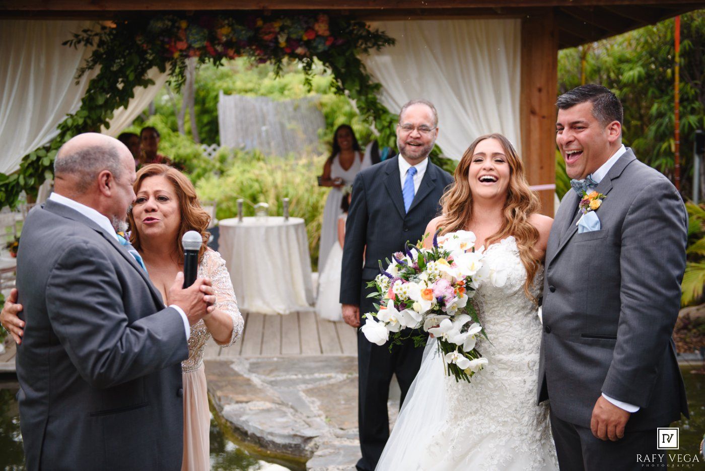 Bride and groom laughing joyfully during outdoor wedding ceremony with floral arch and guests in background.