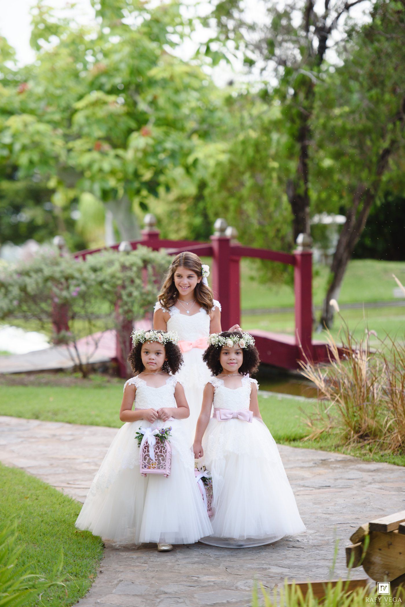 Two flower girls in white tulle dresses with floral crowns walk a garden path with a bridesmaid at an outdoor wedding.