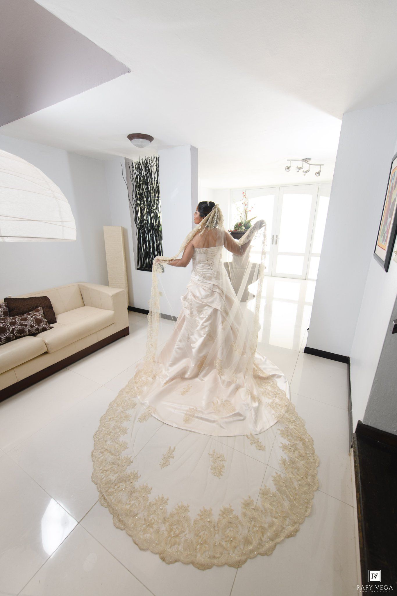 Bride in elegant white gown with gold lace cathedral veil stands in bright modern room before wedding ceremony.
