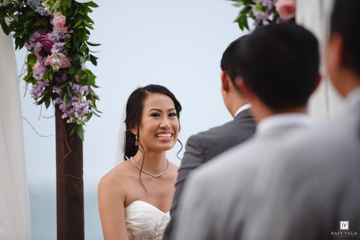 Smiling bride in strapless gown at outdoor wedding ceremony beneath floral arch with purple blooms.