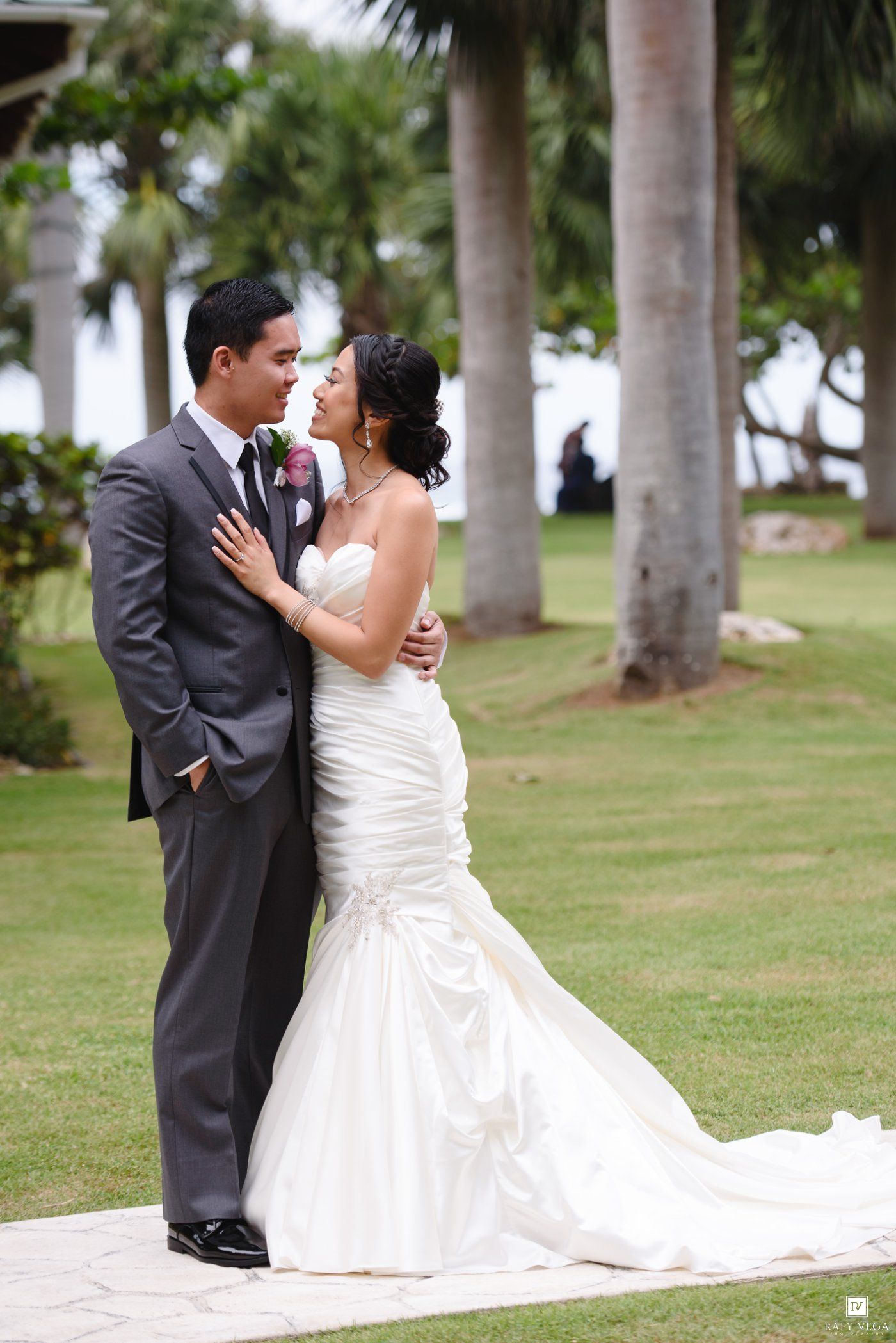 Bride in mermaid gown and groom in gray suit share romantic moment on tropical lawn with palm trees.