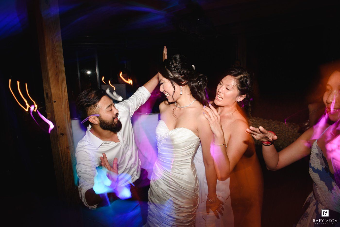 Bride in white strapless gown dancing joyfully with guests under colorful purple and blue lights at a wedding reception.