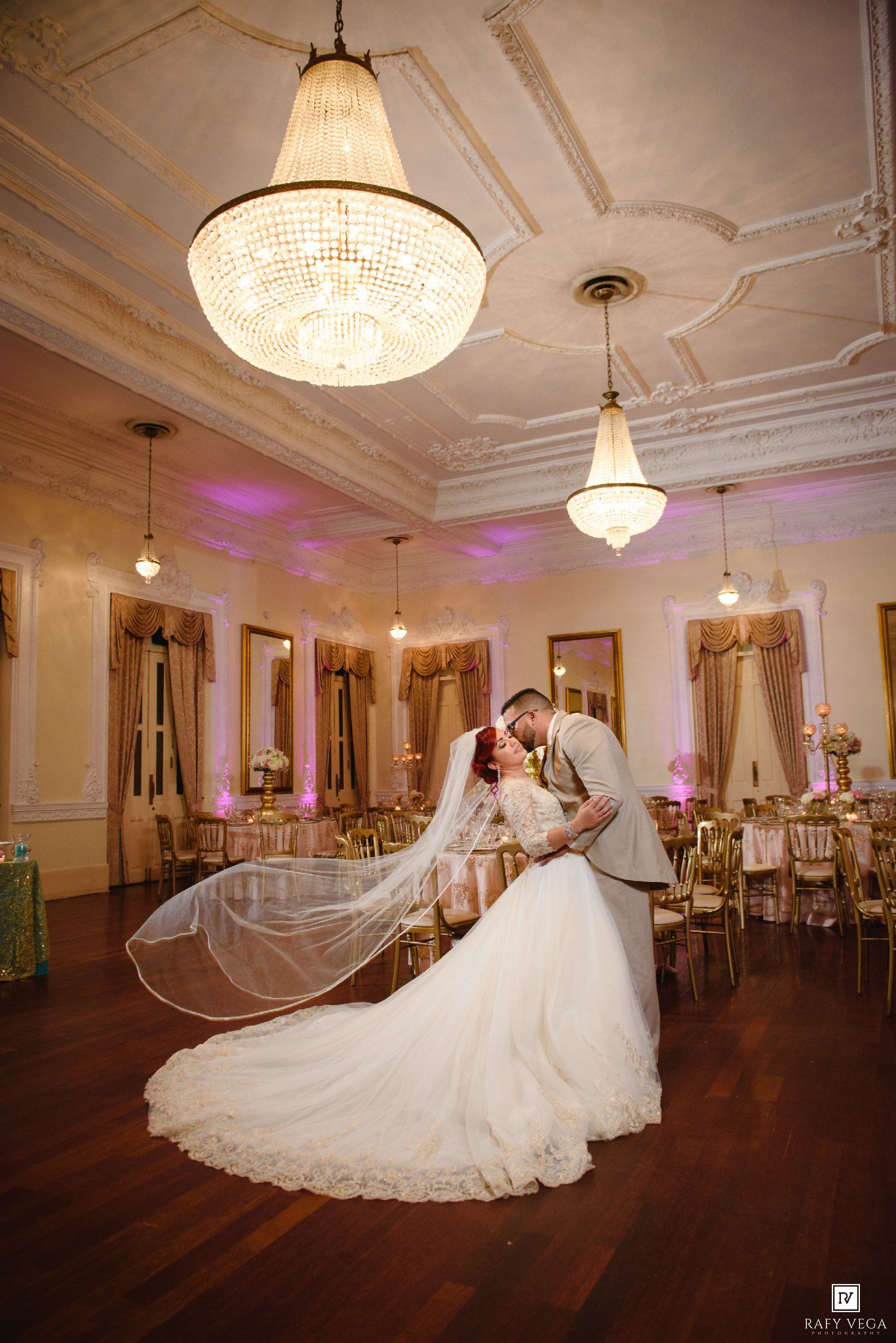 Bride and groom share romantic kiss under grand crystal chandelier in ornate ballroom with flowing veil and pink lighting.