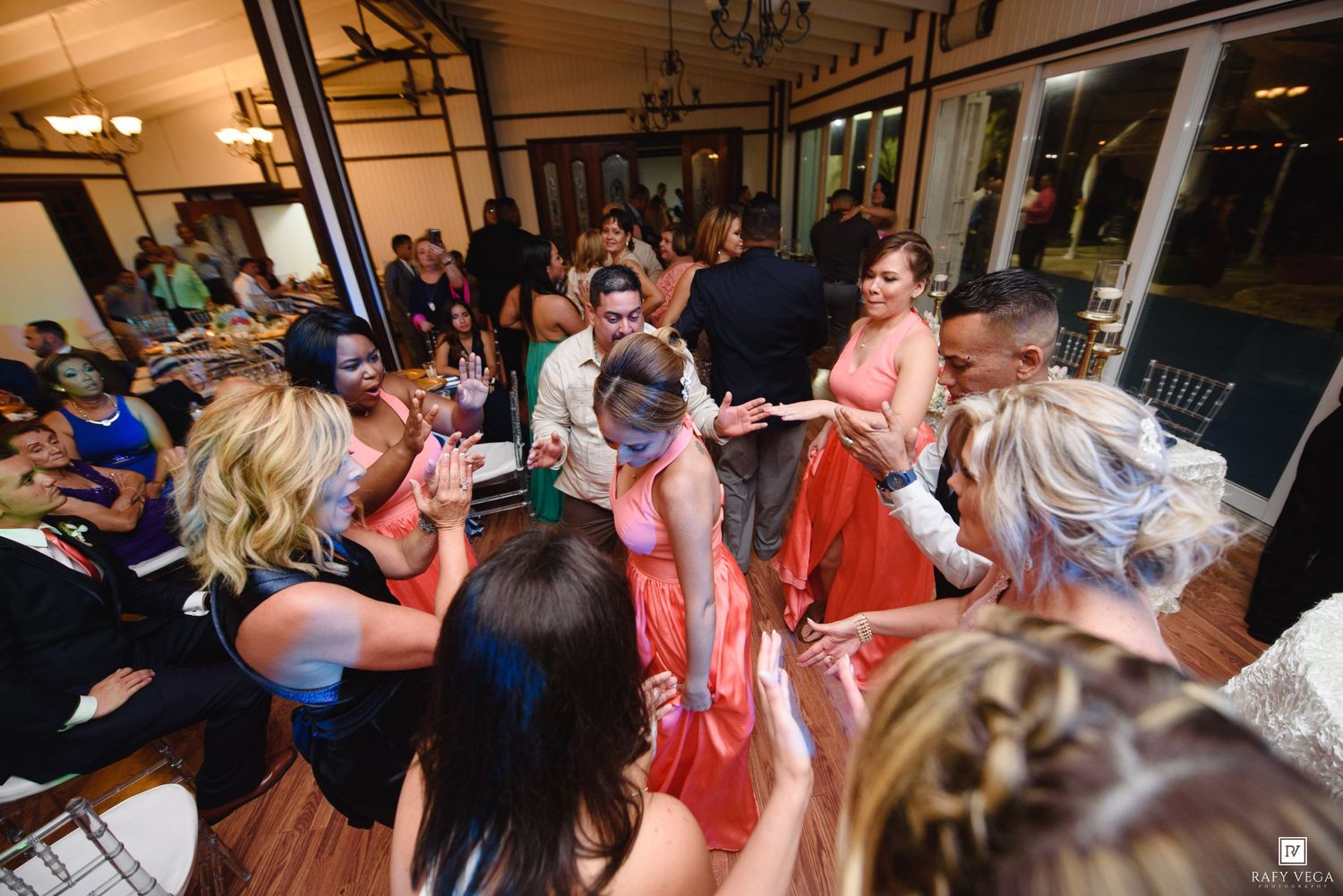Wedding guests dancing together in a lively reception hall, with bridesmaids in coral dresses celebrating on the dance floor.