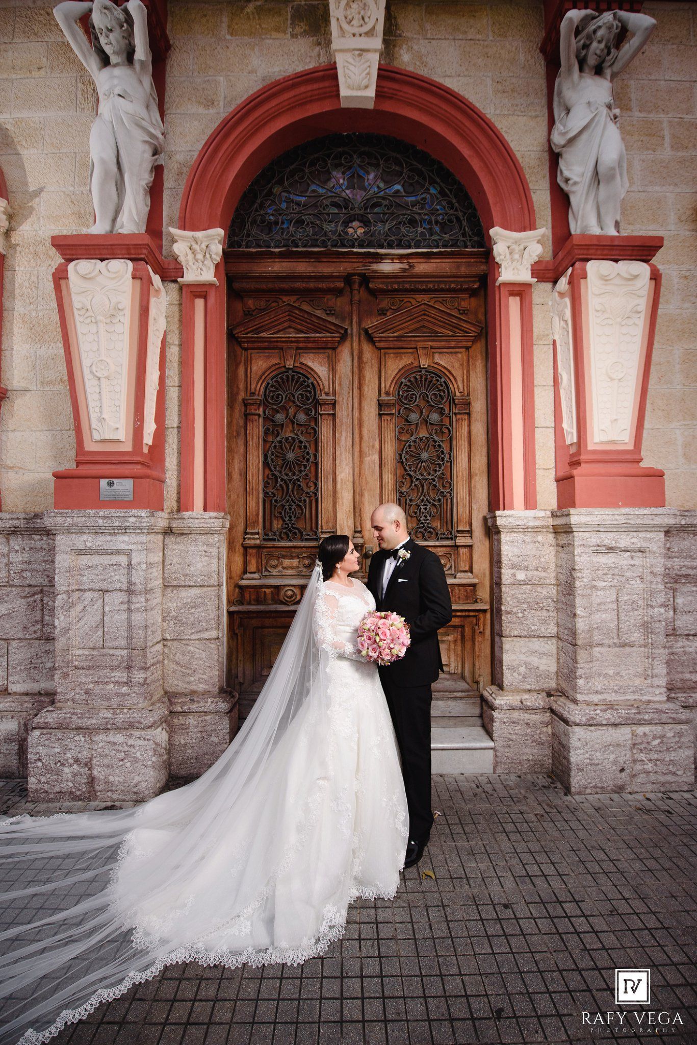 Bride and groom pose before ornate wooden doors with stone statues at an elegant wedding venue.