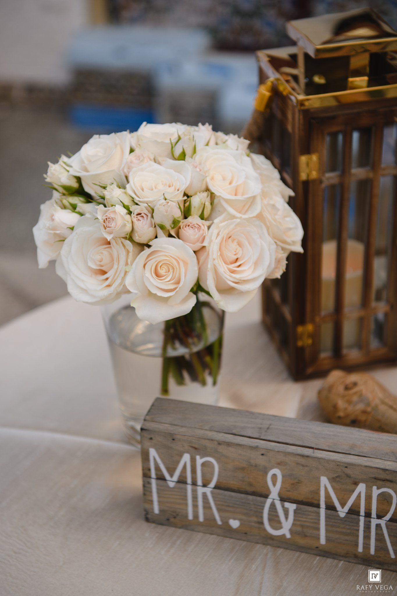 Elegant wedding centerpiece with blush roses in glass vase beside rustic Mr. & Mrs. sign and gold lantern.