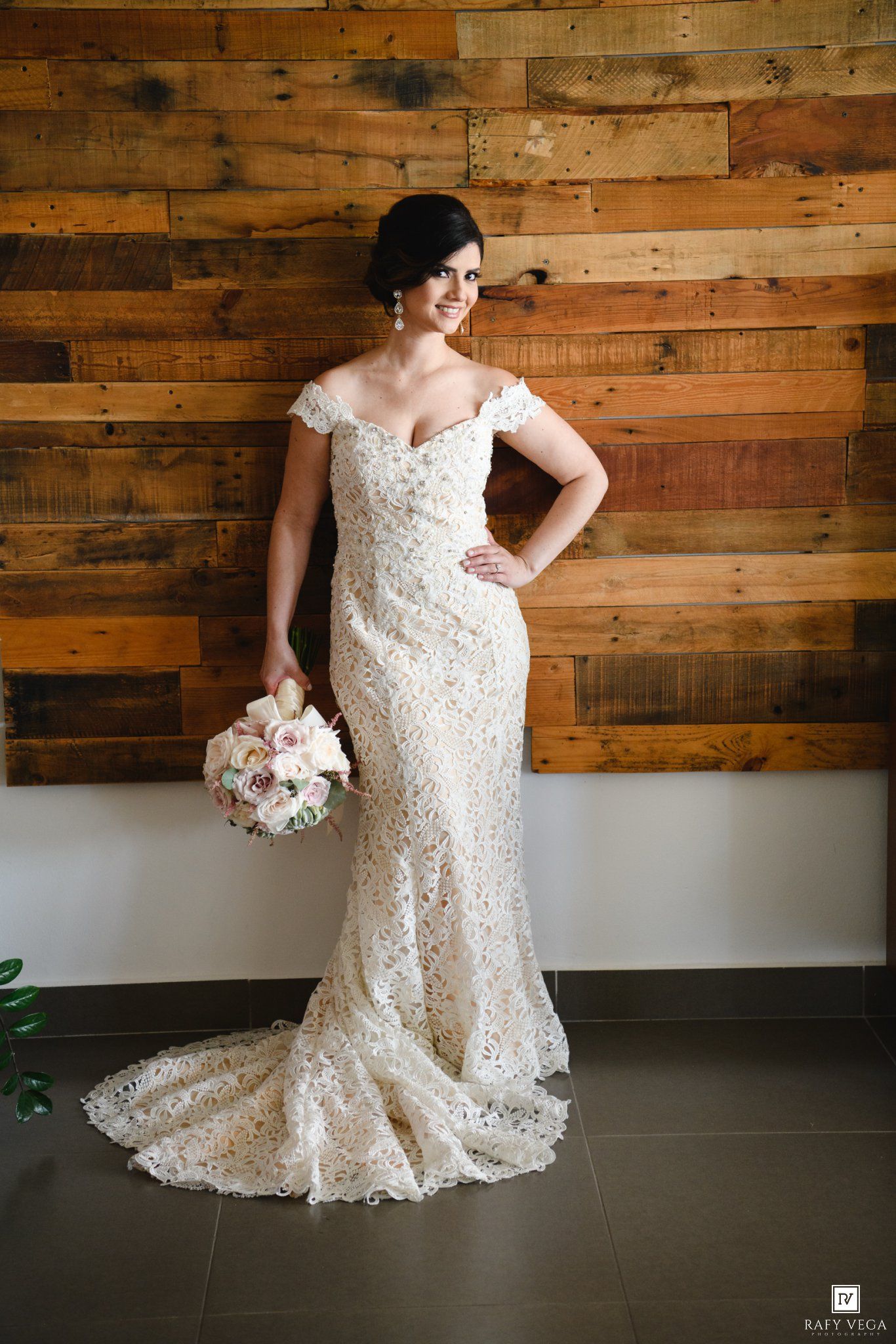 Bride in off-shoulder lace mermaid wedding gown holding pink bouquet, posing before rustic wood plank wall.