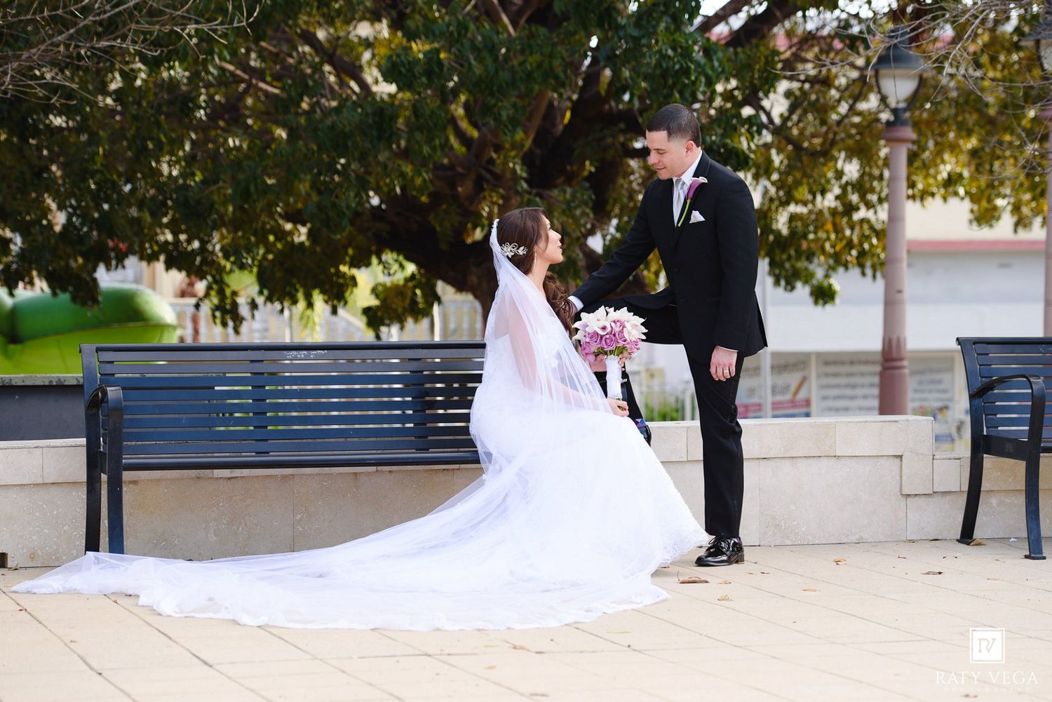 Bride in white gown with veil sits on bench as groom in black suit holds her hand outdoors near trees.