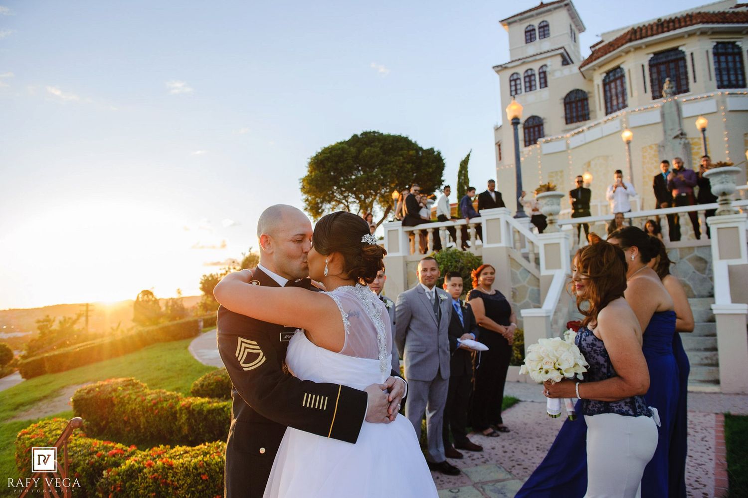 Bride and groom share first kiss at outdoor wedding ceremony near elegant white mansion at golden sunset.