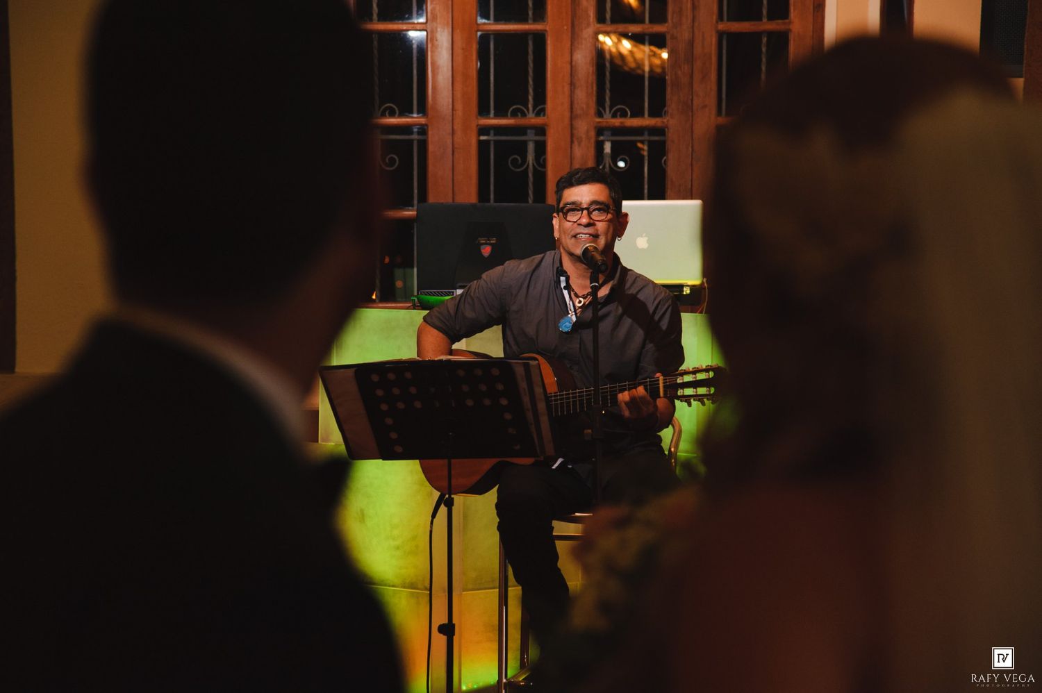 A musician plays acoustic guitar at an elegant indoor wedding reception while guests watch in the foreground.