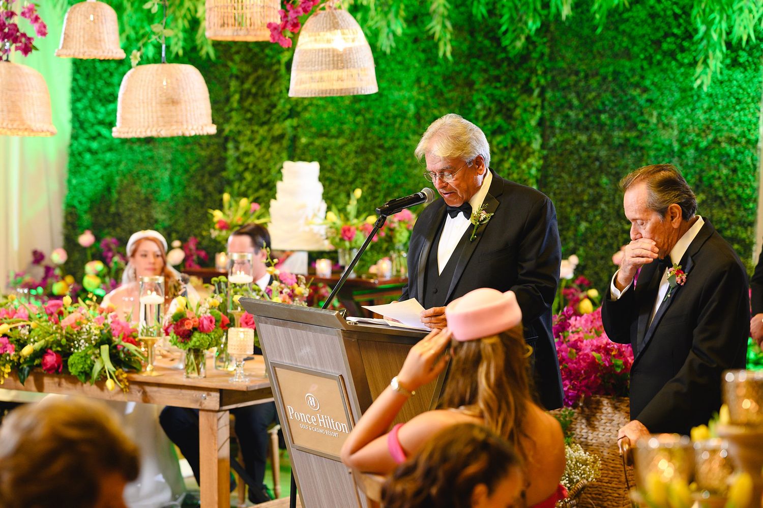 Man in tuxedo giving speech at elegant wedding reception podium with floral arrangements and wedding cake.