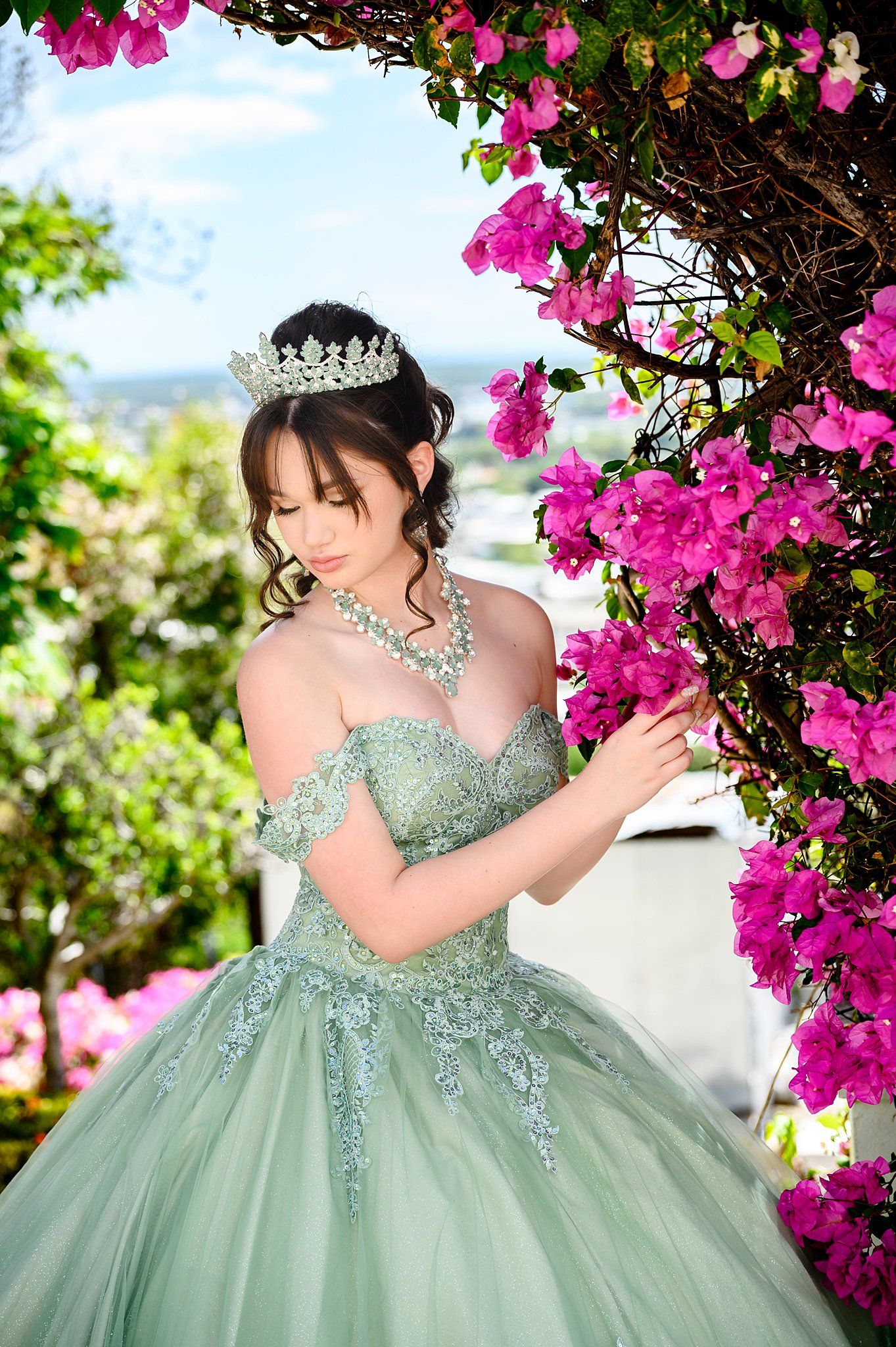 Young woman in sage green quinceañera gown with tiara poses beside vibrant pink bougainvillea flowers outdoors.