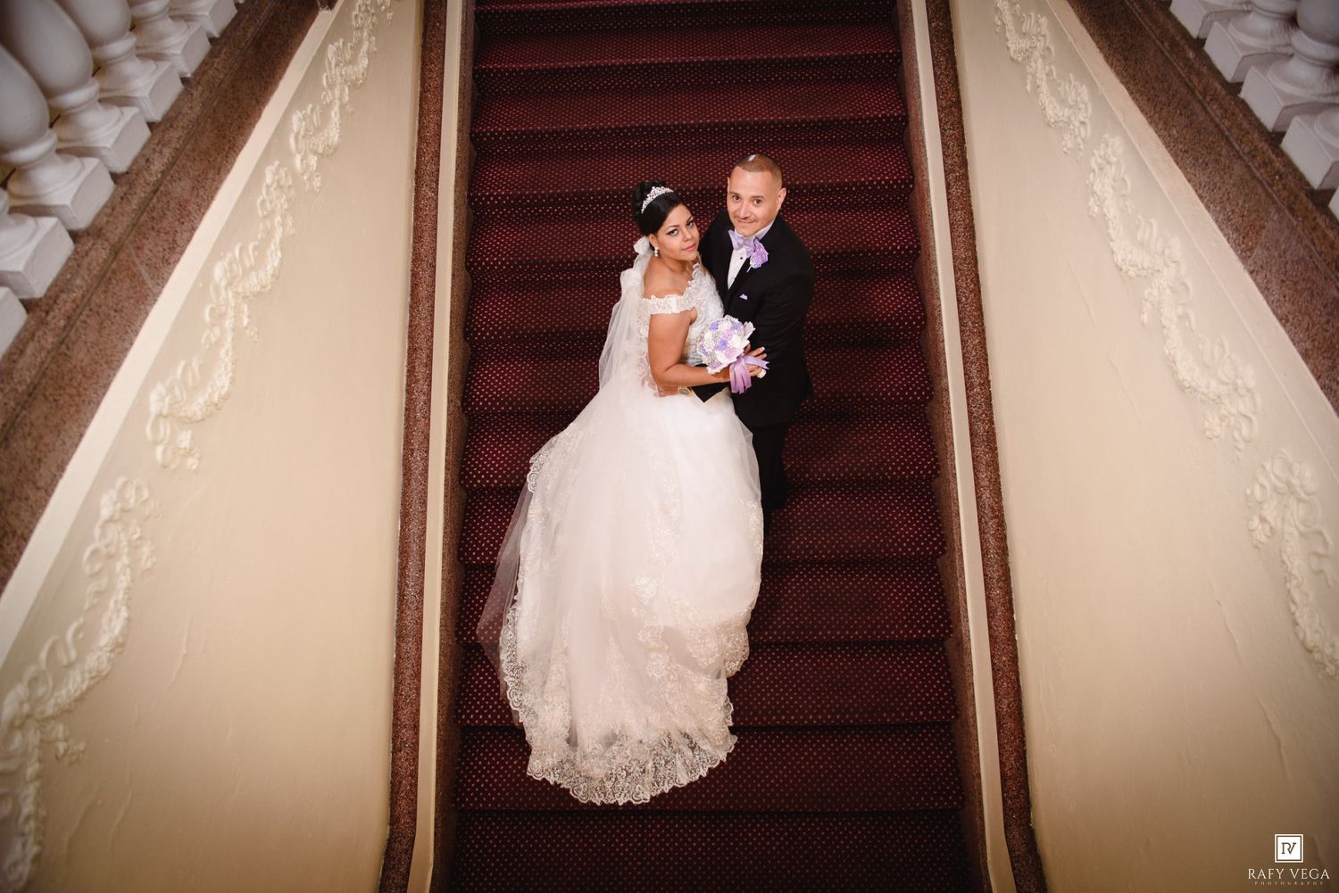 Bride and groom pose elegantly on a grand staircase with red carpet, bride holding a bouquet.