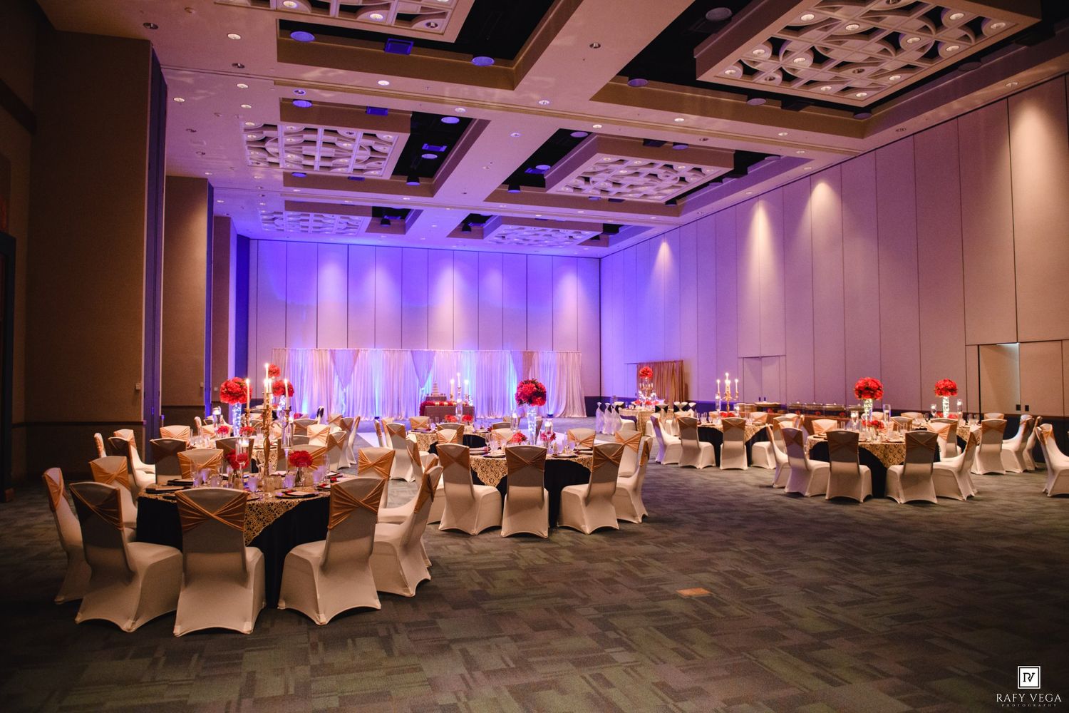 Elegant banquet hall with purple uplighting, white chair covers, gold-trimmed tables, and red floral centerpieces for a wedding reception.