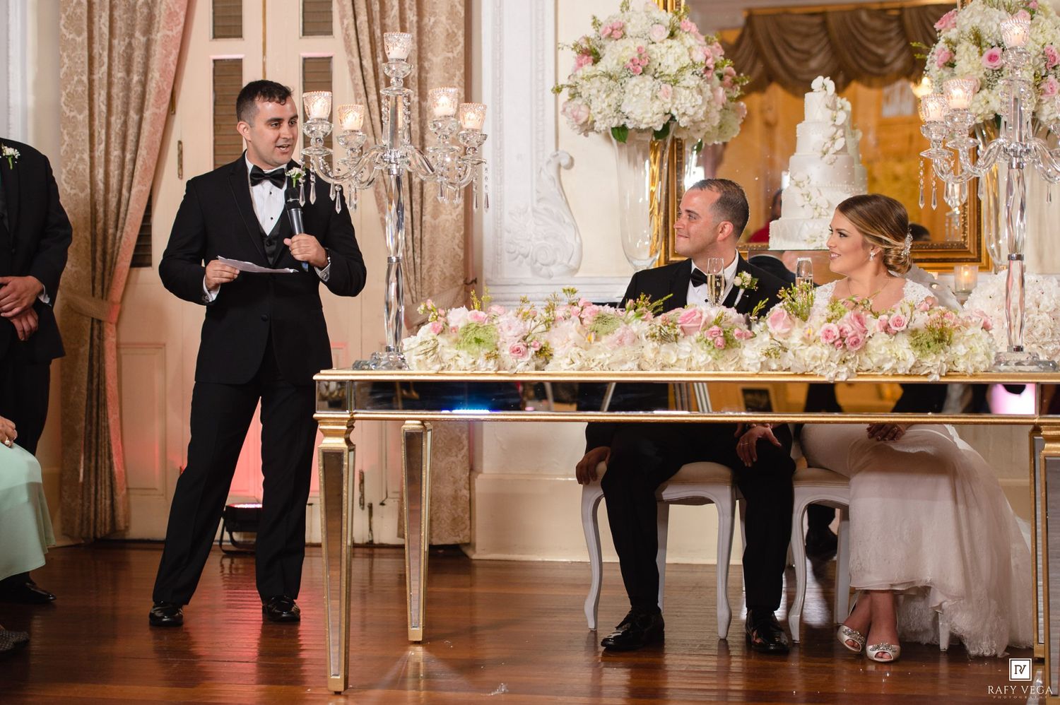 Best man delivers wedding toast at sweetheart table adorned with floral centerpieces.