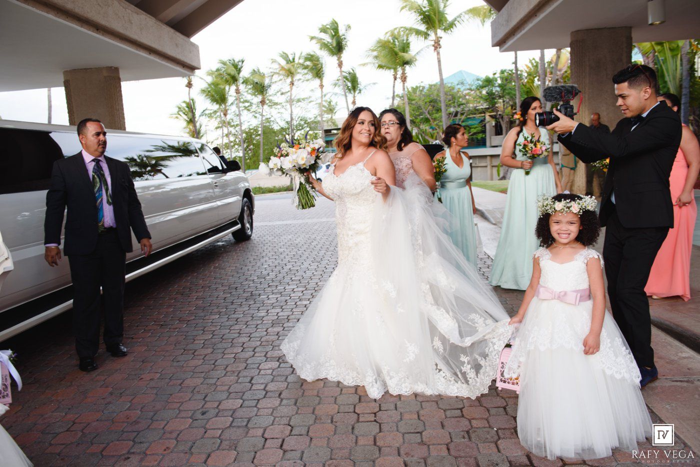 Bride in white lace gown with bridesmaids in mint dresses and flower girl outside venue near white limo on wedding day.