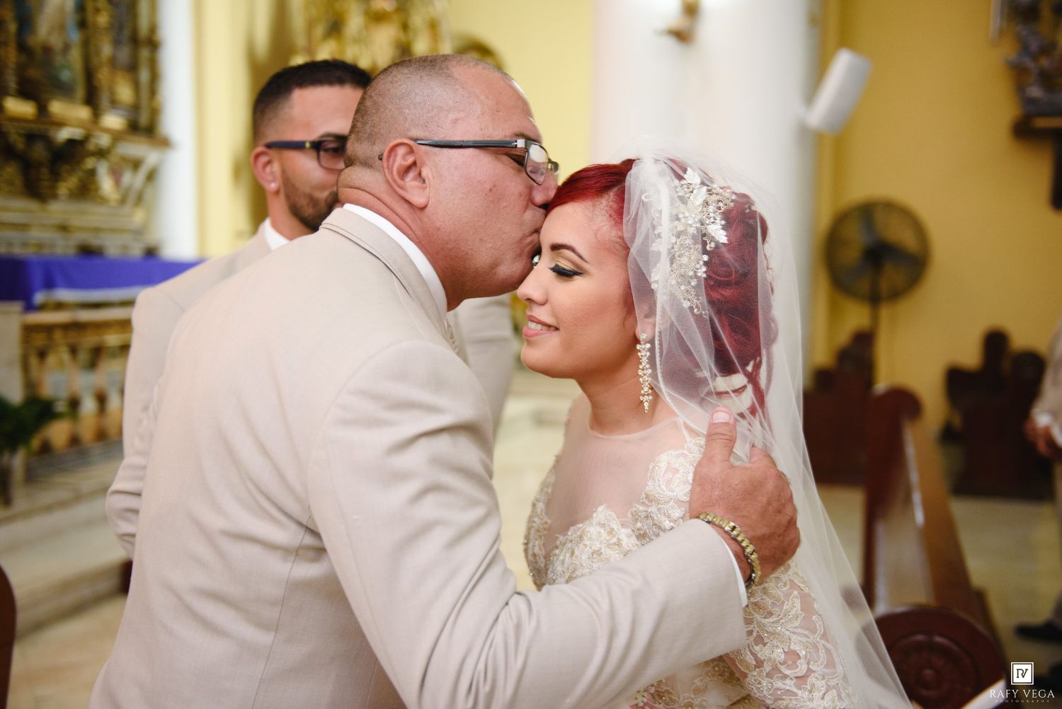 Groom kissing bride on cheek during wedding ceremony in church, bride wearing lace gown and veil.