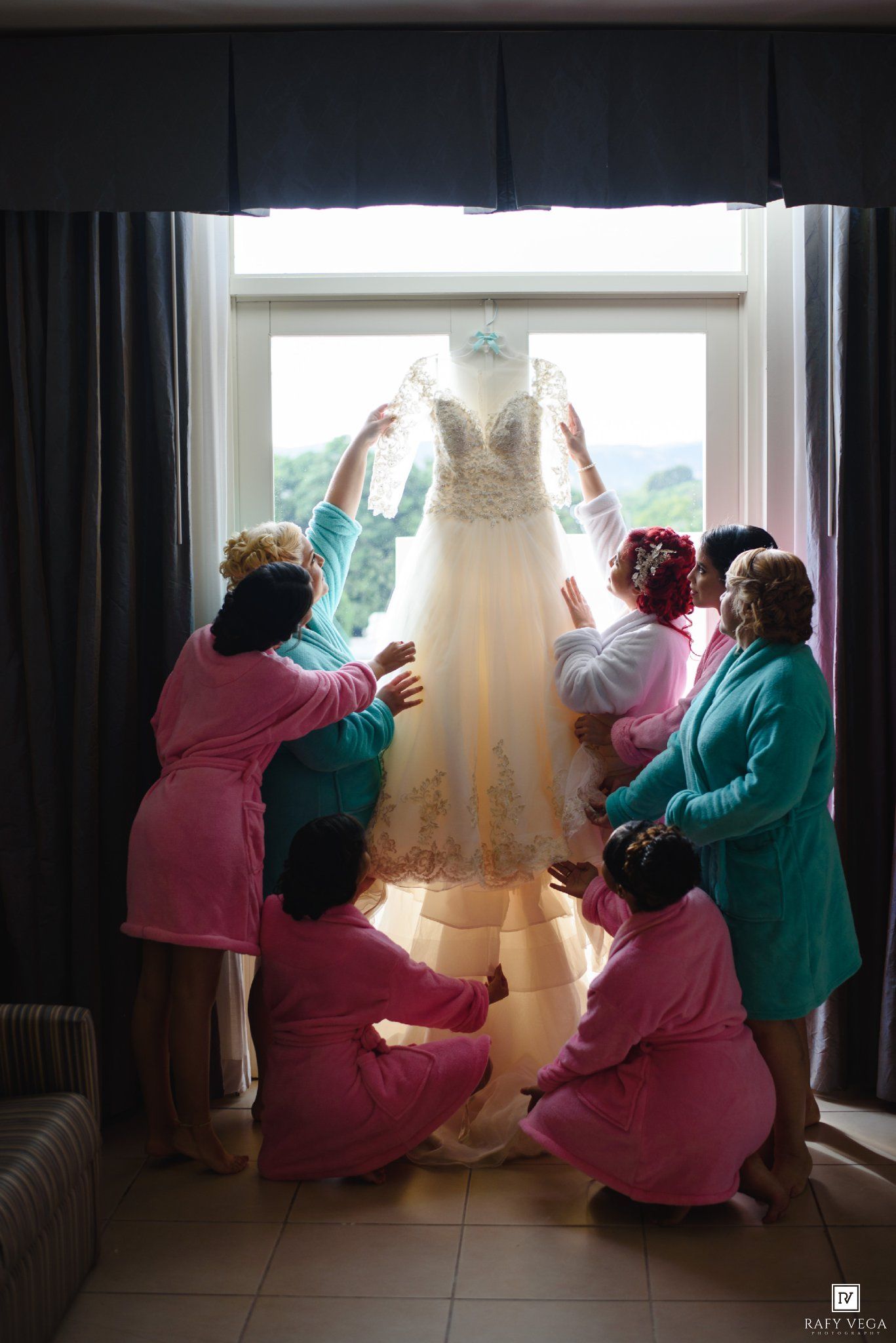 Bridesmaids in pink and teal robes admire a lace wedding dress hanging in a sunlit window.