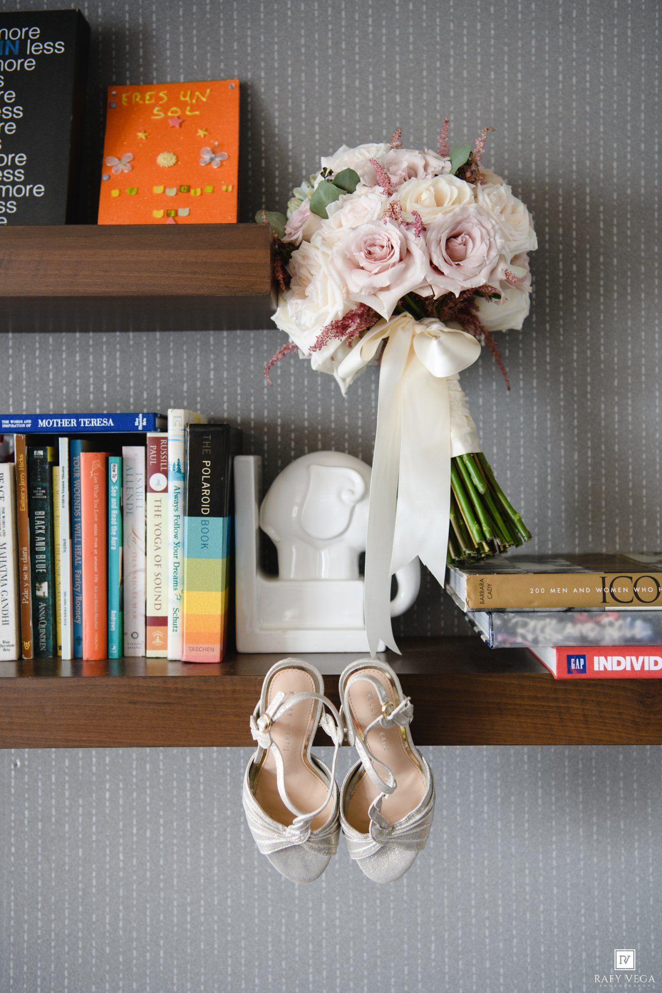 Bridal bouquet of blush pink roses with white ribbon resting on wooden shelf beside white heels and colorful books.