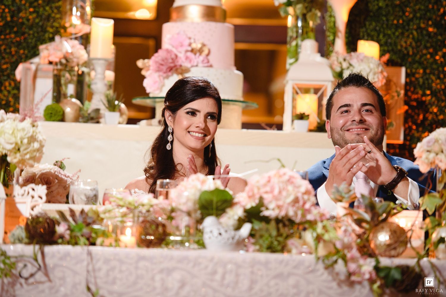 Smiling bride and groom seated at elegantly decorated wedding reception table with floral arrangements and tiered cake.