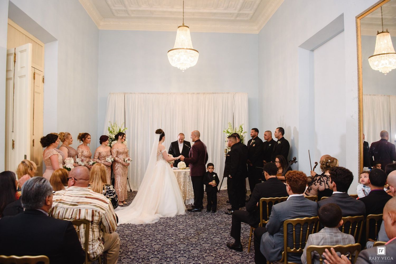 Wedding ceremony in elegant blue room with bridesmaids, groomsmen, and guests seated around couple.