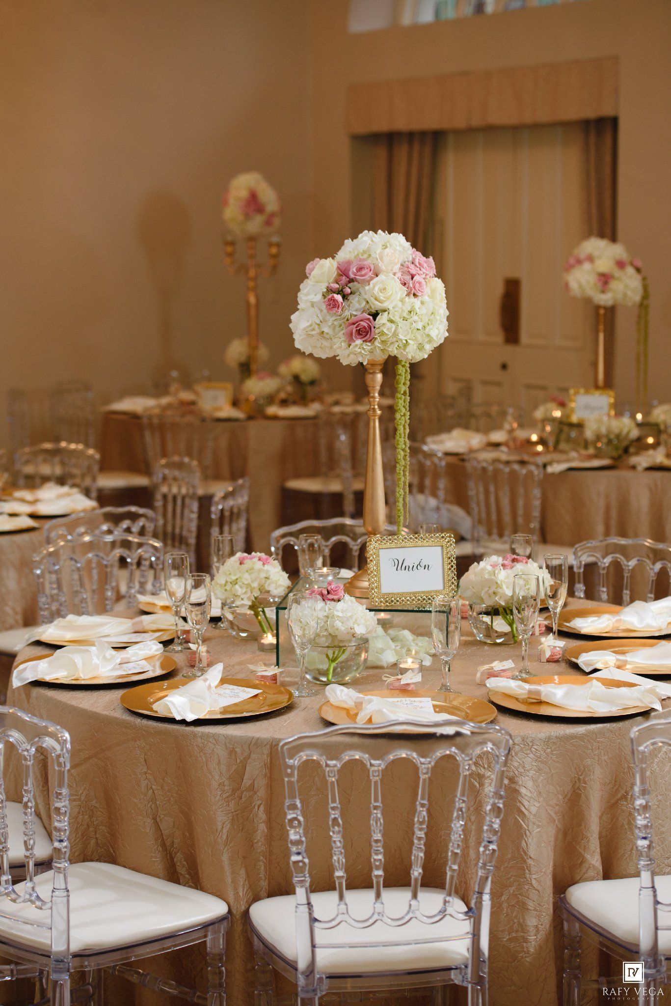 Elegant wedding reception table with gold linens, crystal chairs, and tall floral centerpiece of white and pink roses.