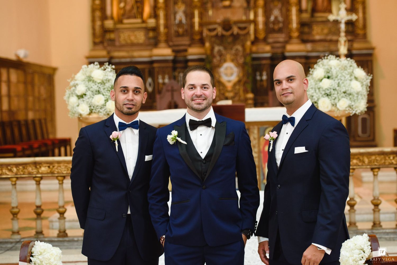 Three groomsmen in navy blue tuxedos with bow ties pose inside an elegant church with floral arrangements.