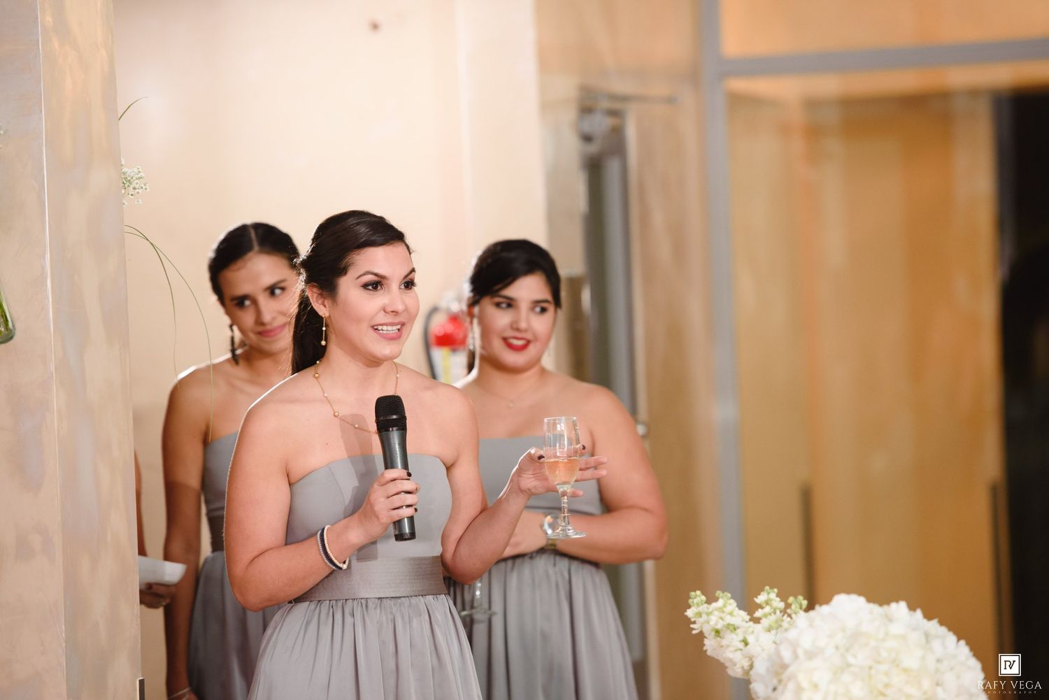 Three bridesmaids in gray dresses delivering a speech at a wedding reception, one holding a microphone.