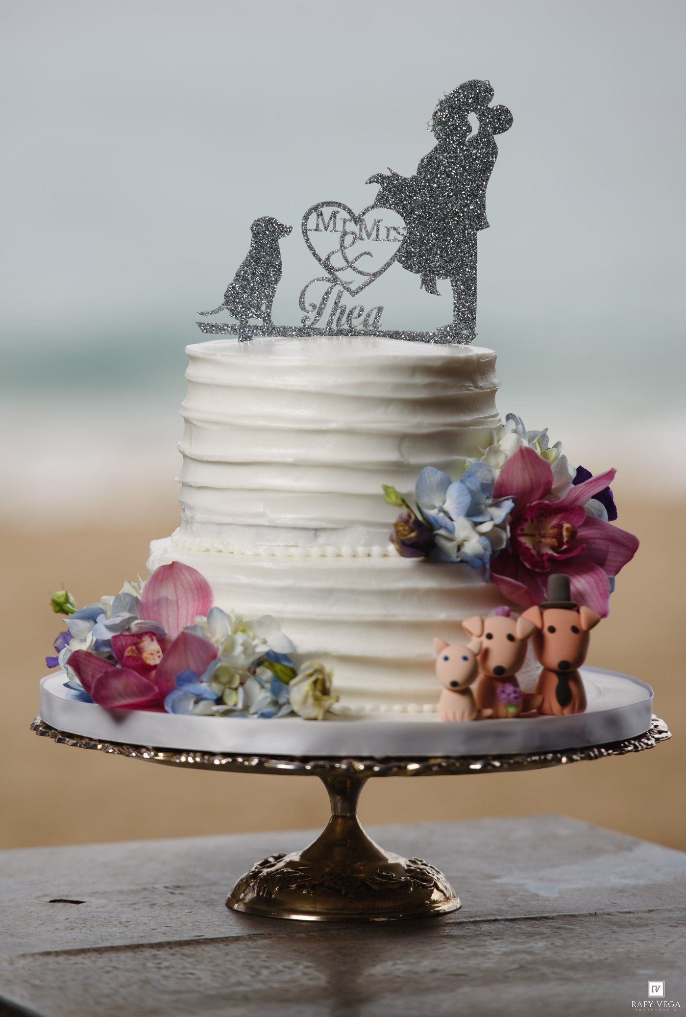 Two-tier white wedding cake topped with couple and dog silhouette, adorned with orchids and pig figurines.