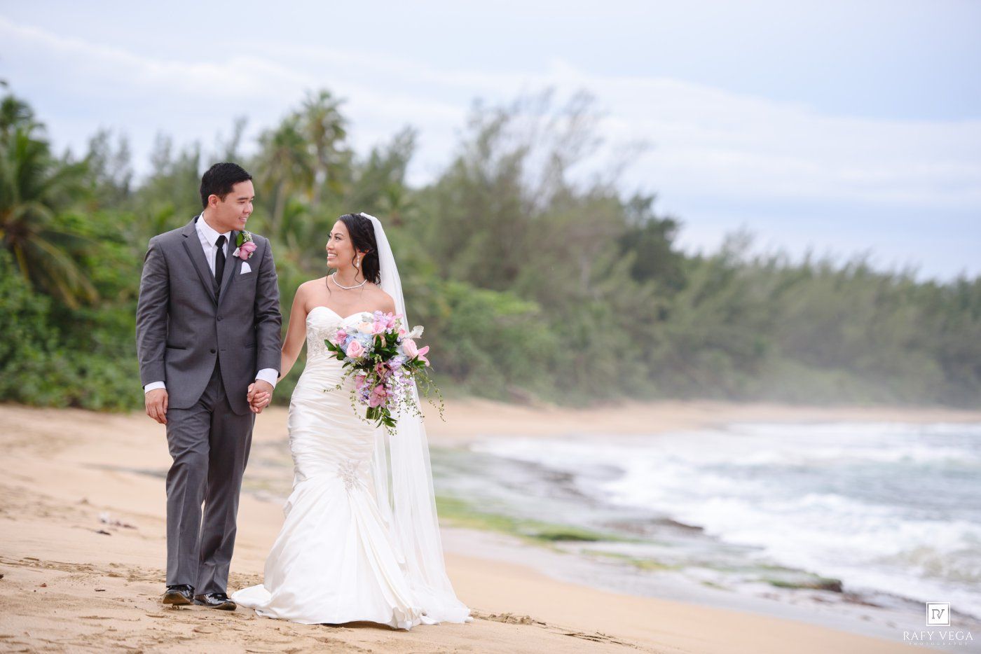 Bride and groom walking hand in hand on a tropical beach, bride holding a pink bouquet in a white gown.