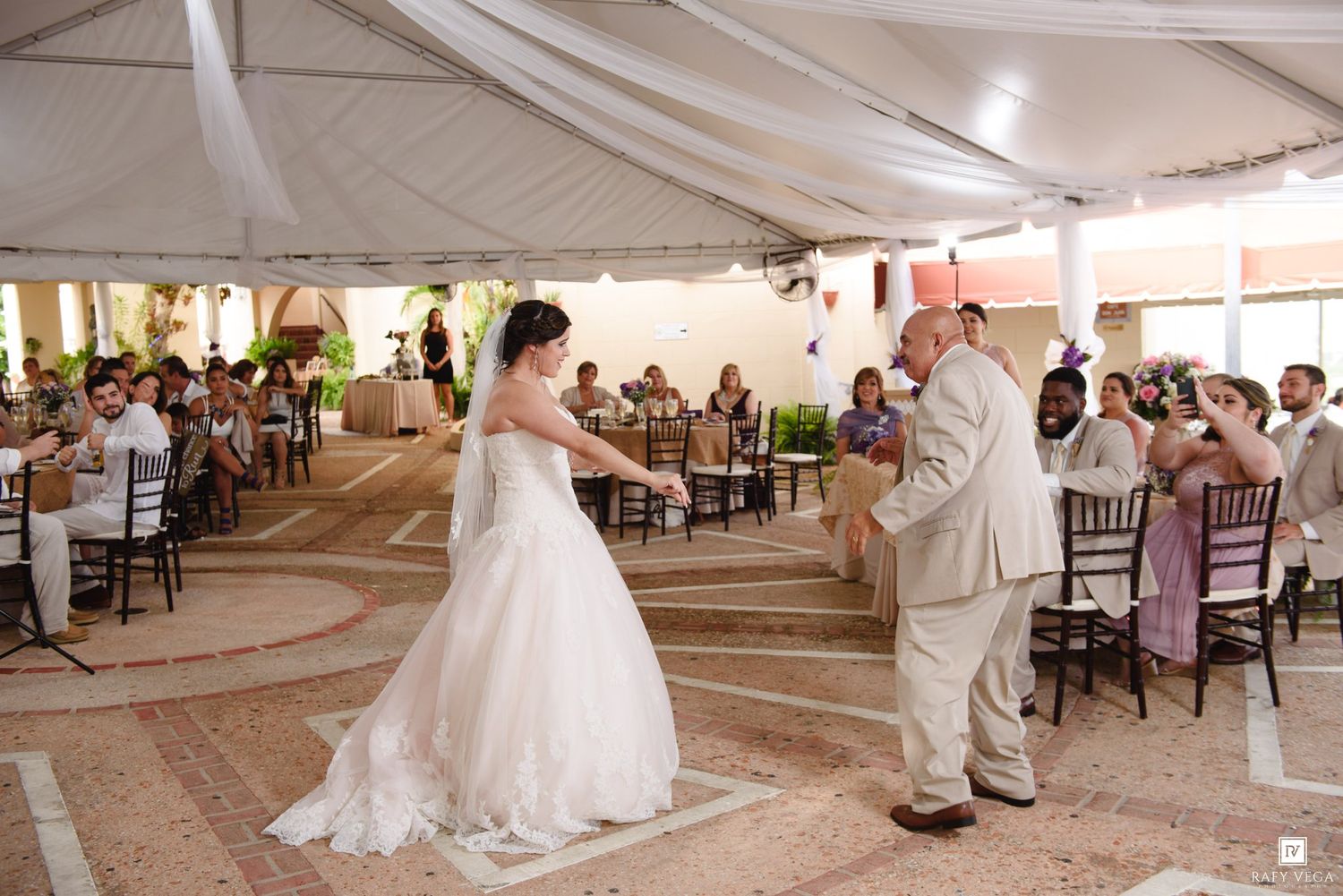 Bride and groom share their first dance under a white tent at an outdoor wedding reception with guests seated around them.