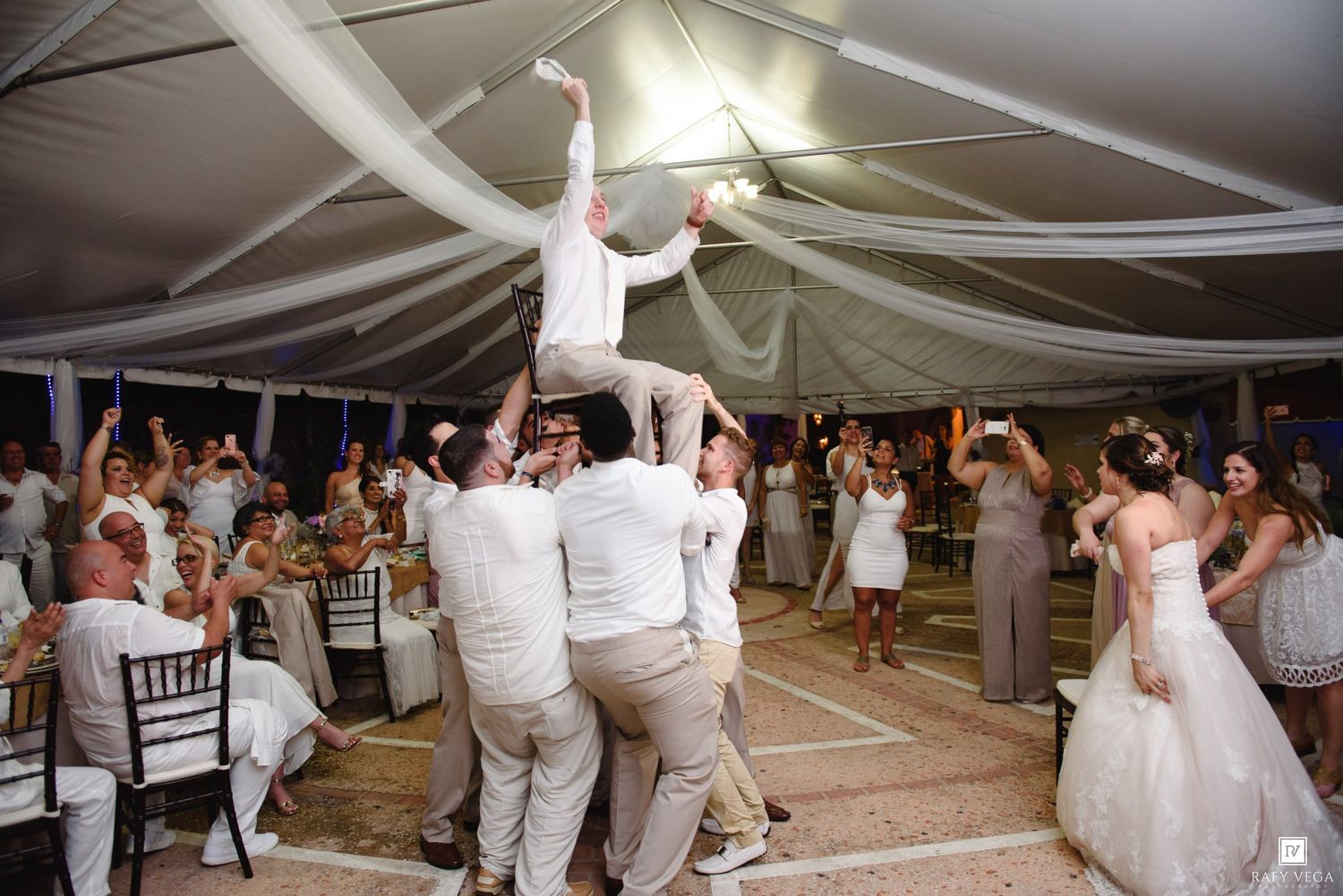 Groom lifted by groomsmen during lively outdoor wedding reception under white draped tent.
