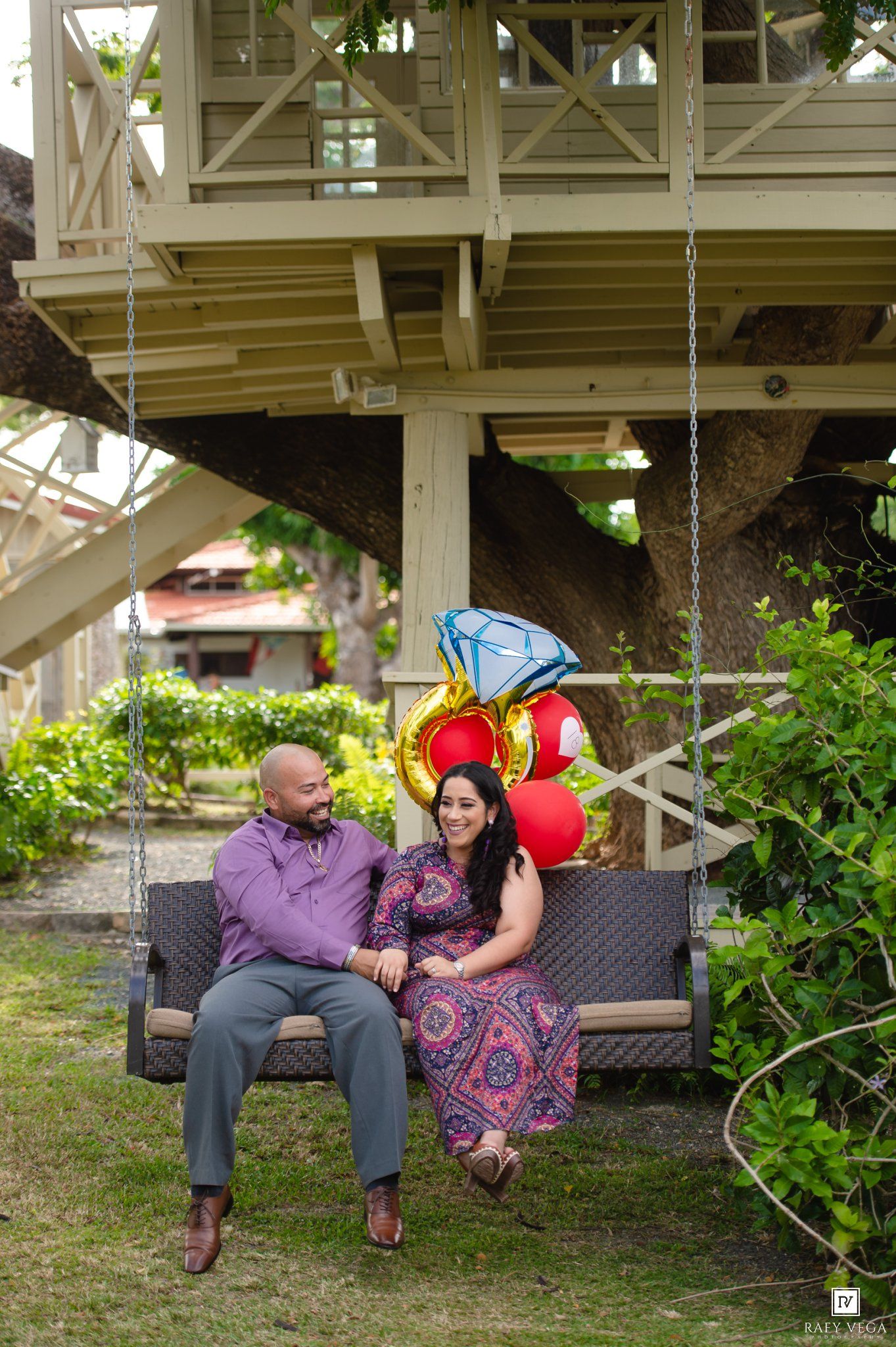 Couple sitting on a wooden swing beneath a treehouse with engagement ring balloons during an outdoor proposal photo session.