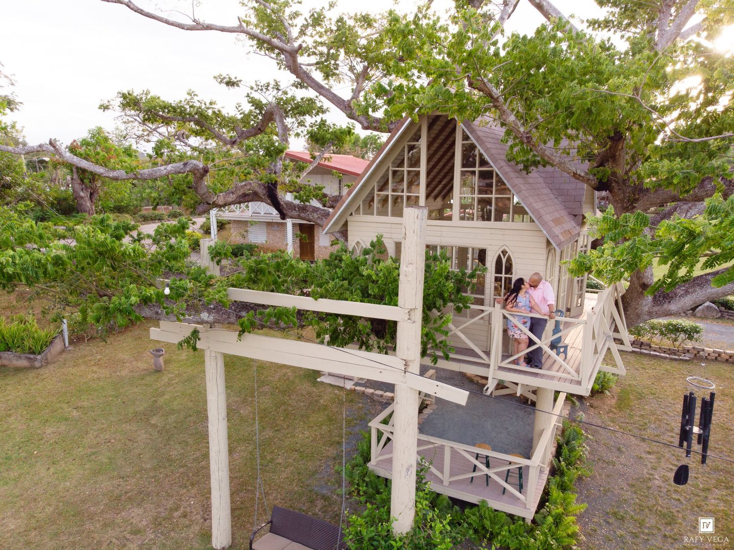 Aerial view of a charming A-frame treehouse with a wooden deck, surrounded by lush green trees and tropical landscaping.