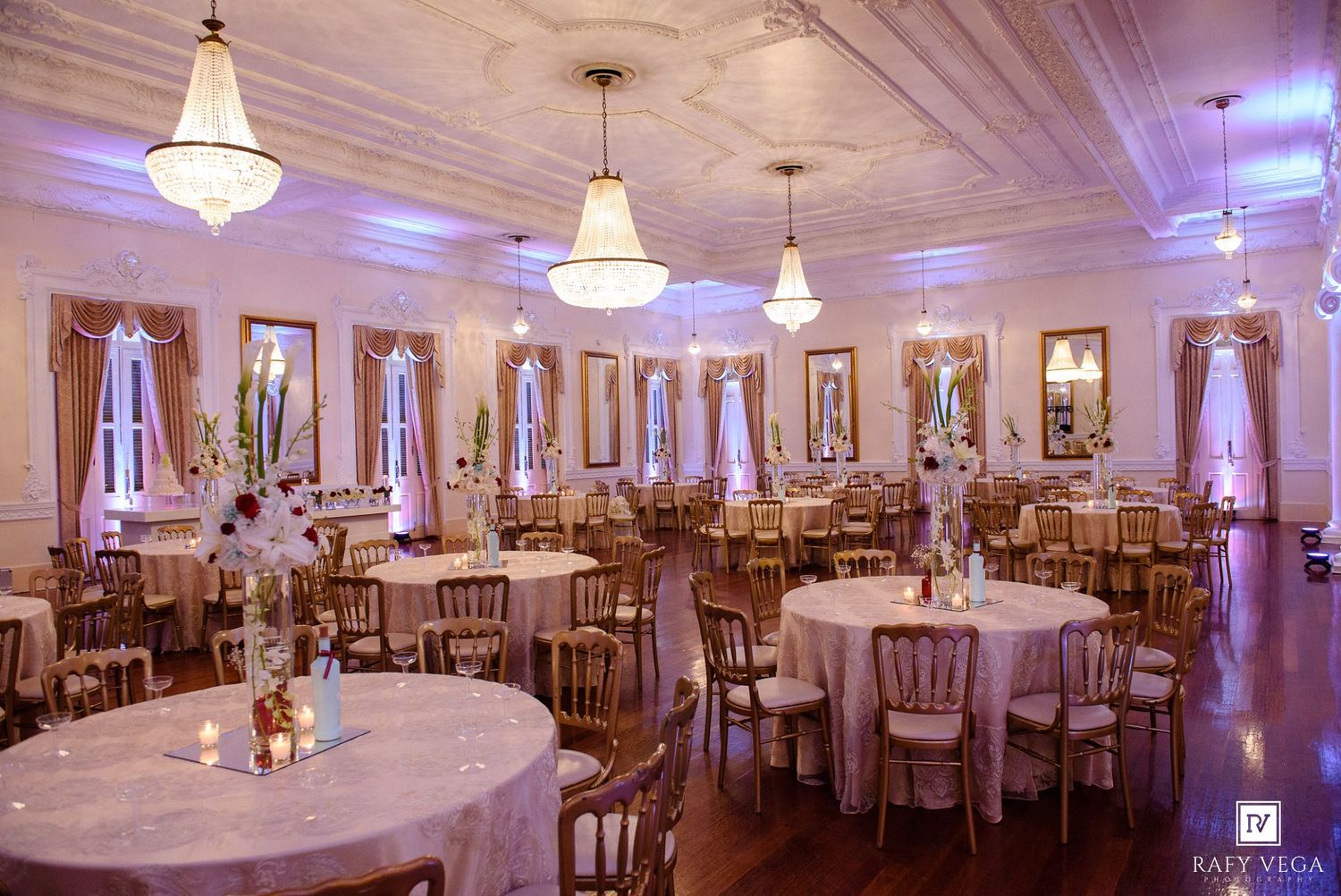 Elegant ballroom with round tables, gold chairs, crystal chandeliers, and soft purple uplighting for a wedding reception.