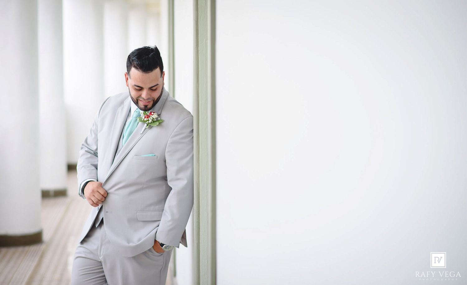 Smiling groom in light gray suit with mint tie and boutonniere leaning against white wall outdoors.