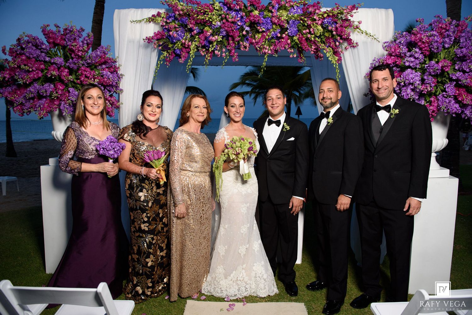 Wedding party poses under floral arch with purple blooms, bride in white gown with groom and guests in formal attire outdoors.
