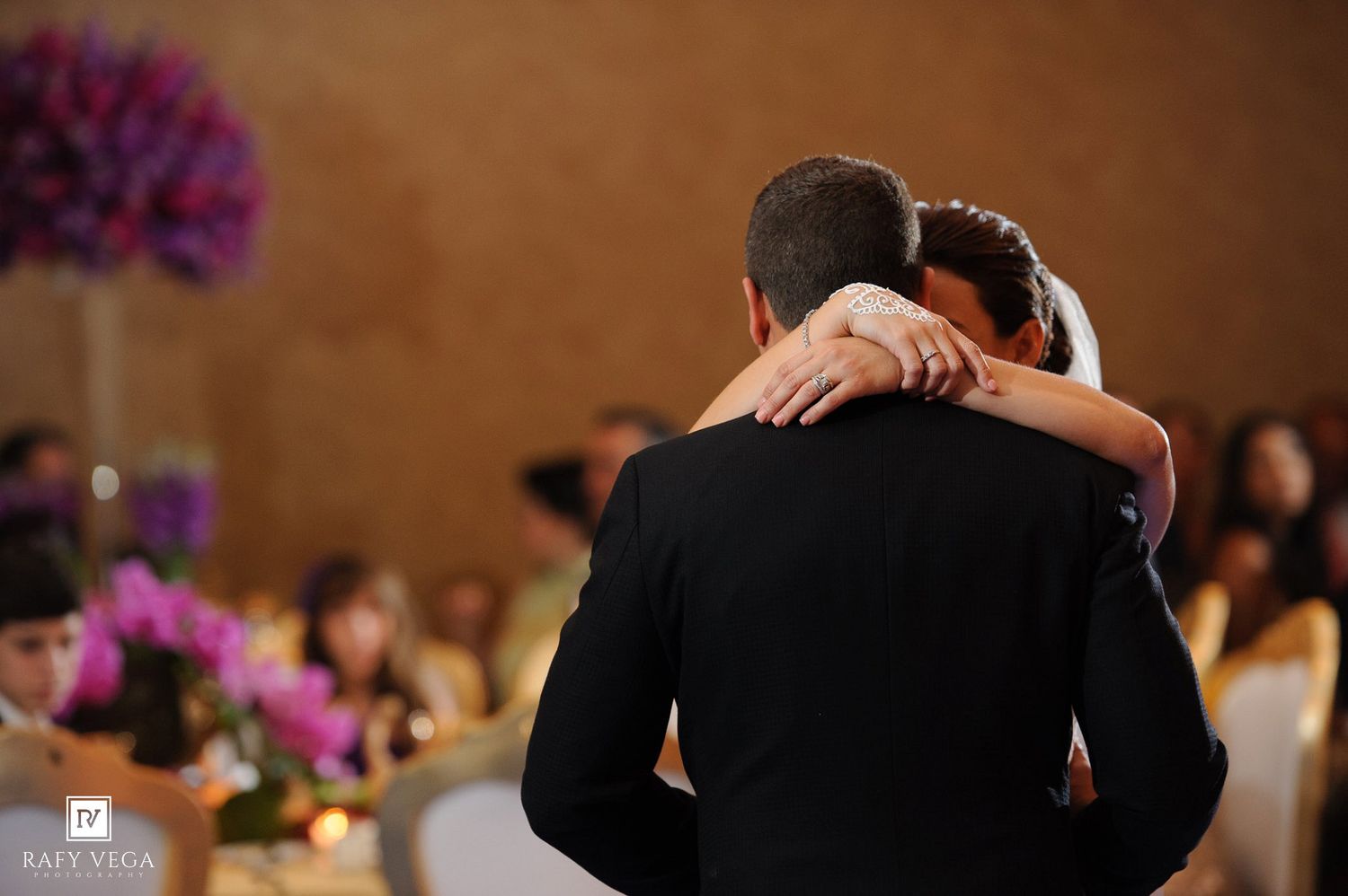 Bride and groom share first dance embrace at elegant wedding reception with purple floral centerpieces.